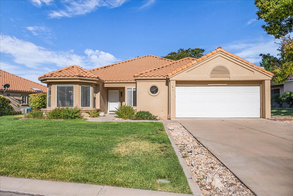 Stucco exterior with a terracotta tile roof, featuring a bay window with multiple panes, an attached garage with a white overhead door, a circular window accent, and a concrete driveway bordered by a rock bed