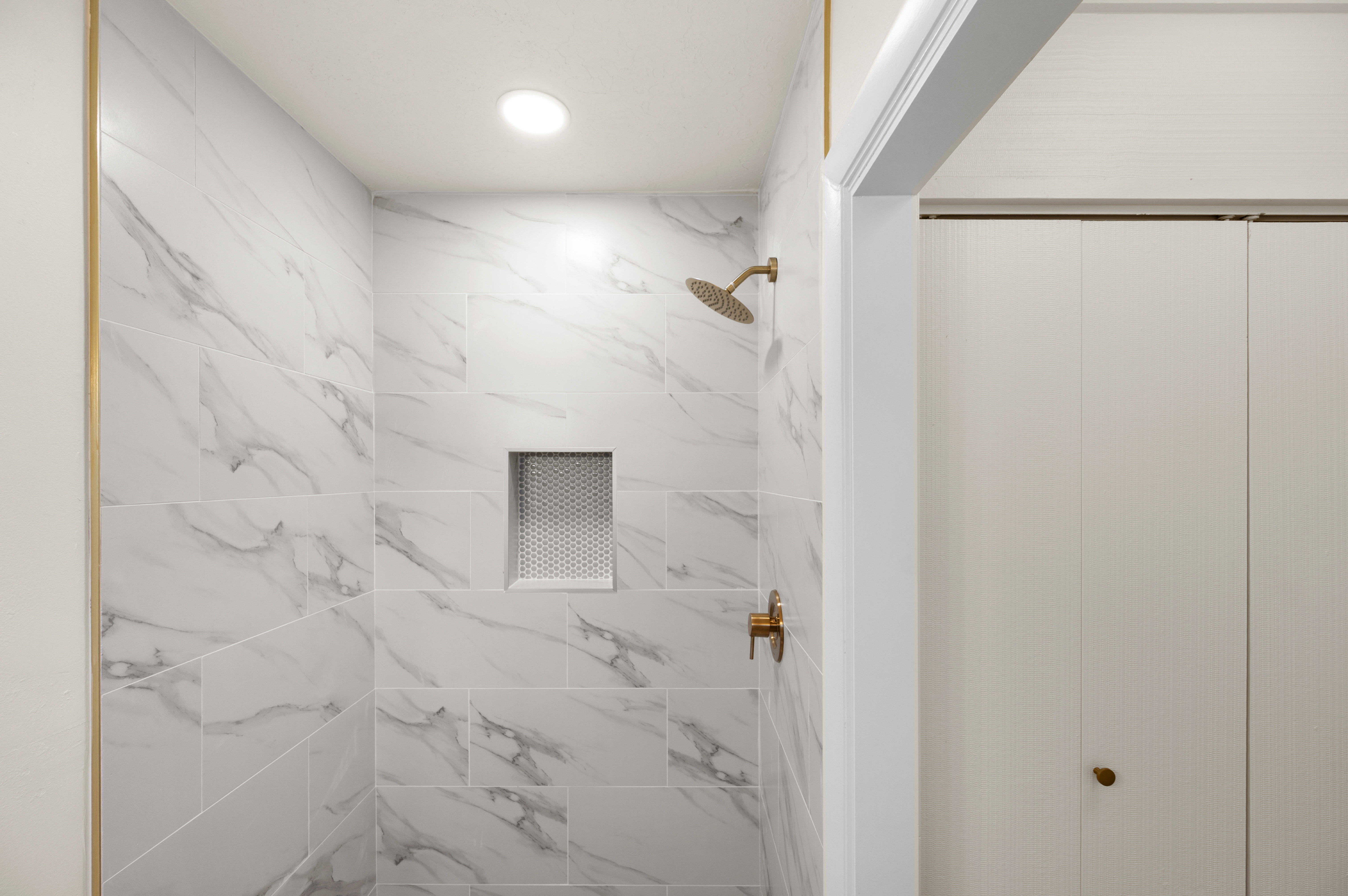 Shower with white marble-patterned tile, gold-finish fixtures, recessed lighting, and a built-in shower niche with a textured back wall