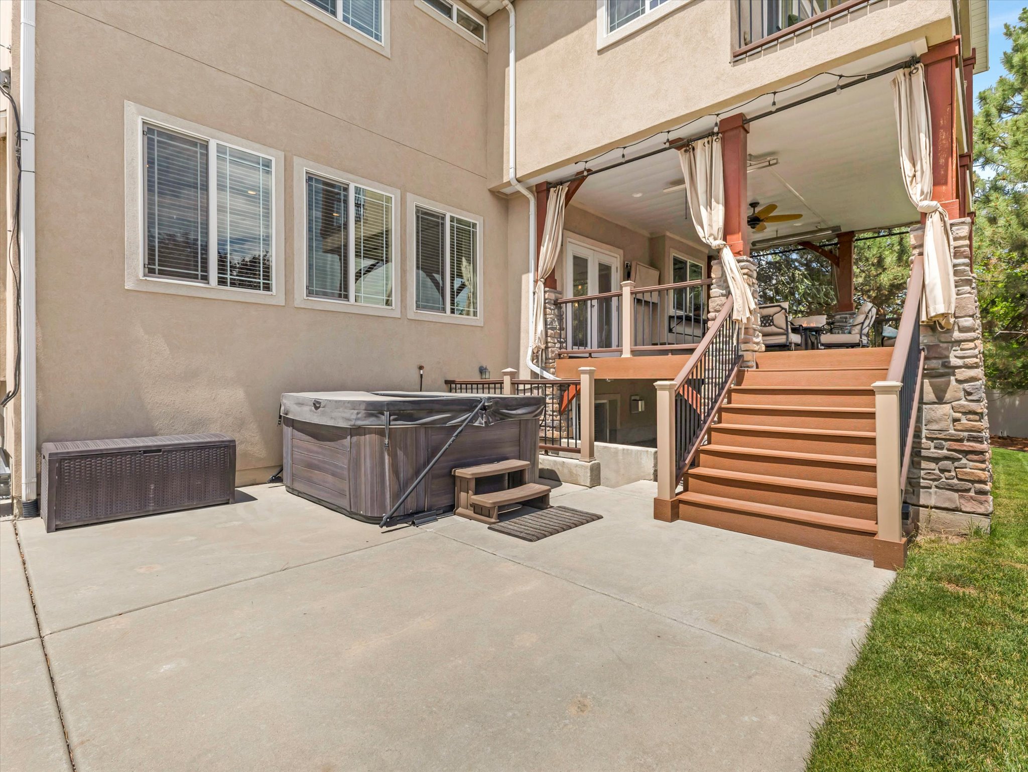 Expansive concrete patio featuring a hot tub, leading to a covered upper deck with built-in lighting, a ceiling fan, and privacy curtains