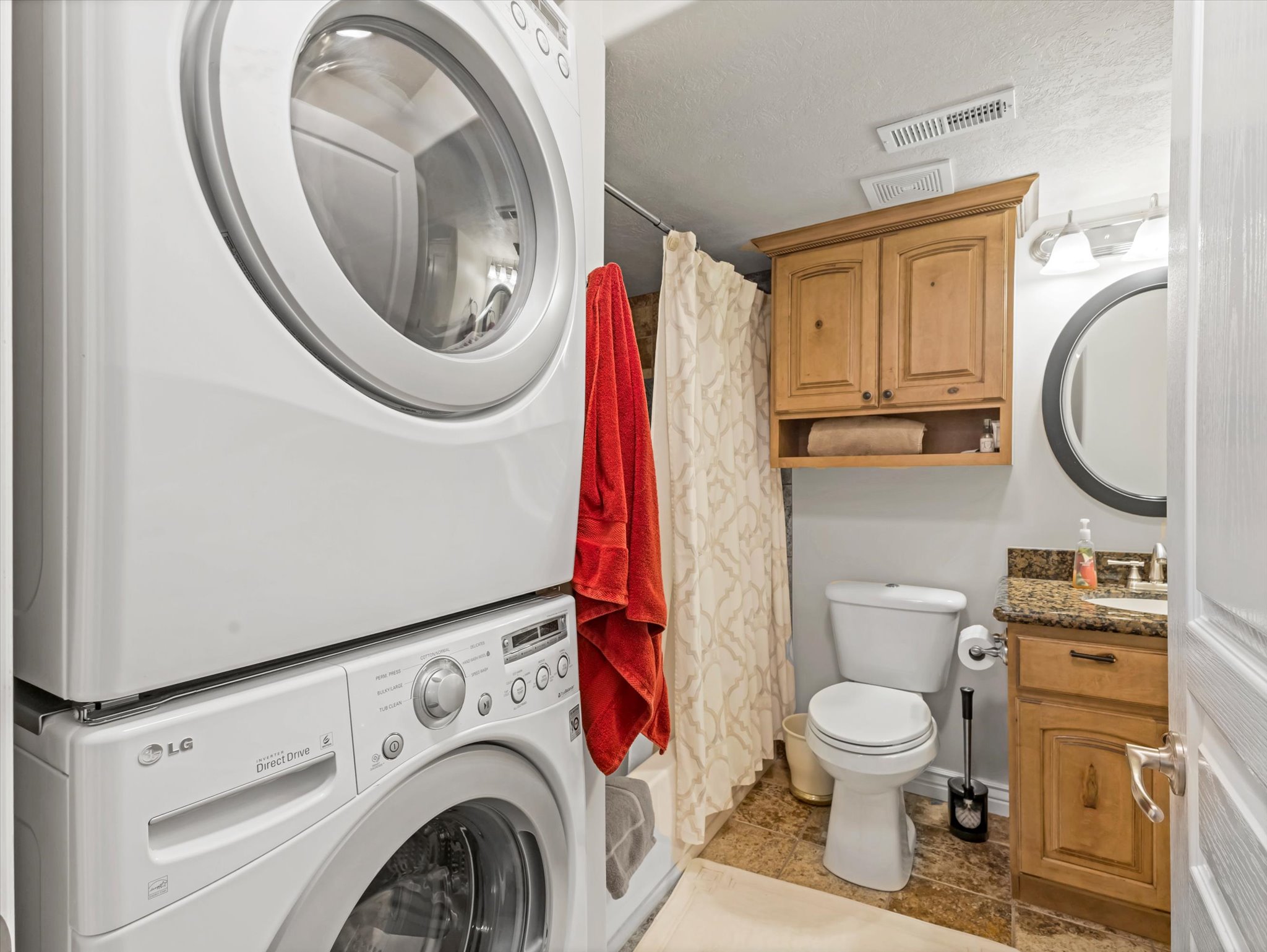Basement laundry area with stacked washer and dryer, featuring a nearby bathroom with a granite-style countertop, wood-finish vanity, and overhead wood-finish cabinetry