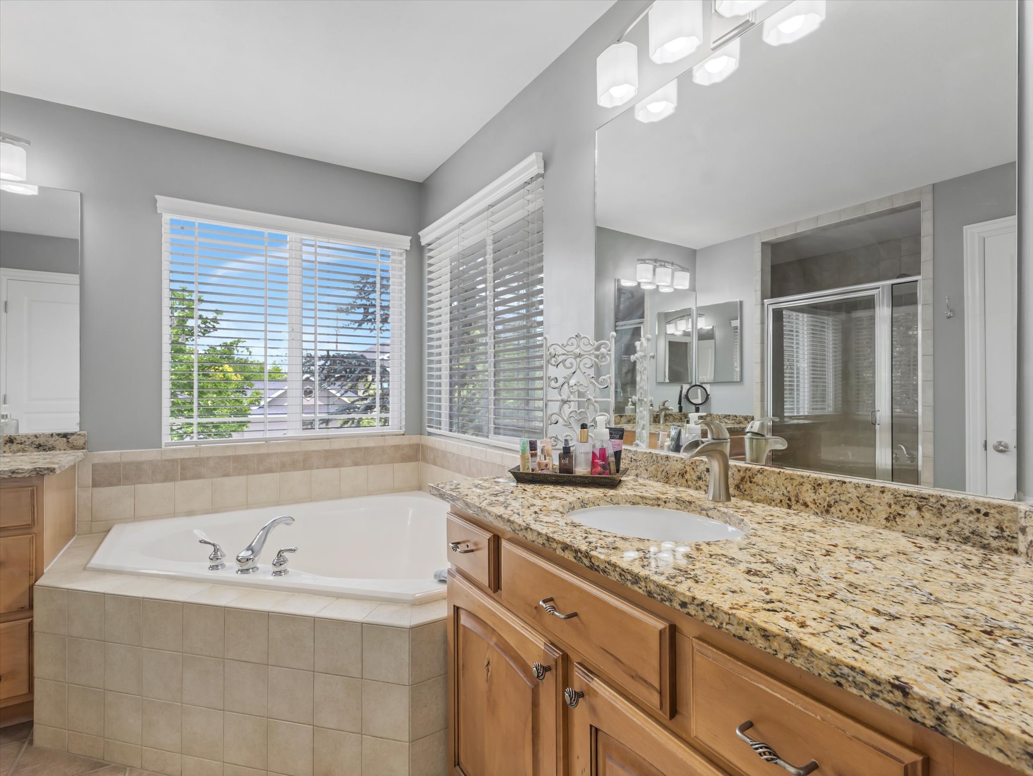 Primary suite bathroom featuring a built-in soaking tub, dual windows with blinds, a spacious vanity with a granite-style countertop, and an expansive wall-mounted mirror