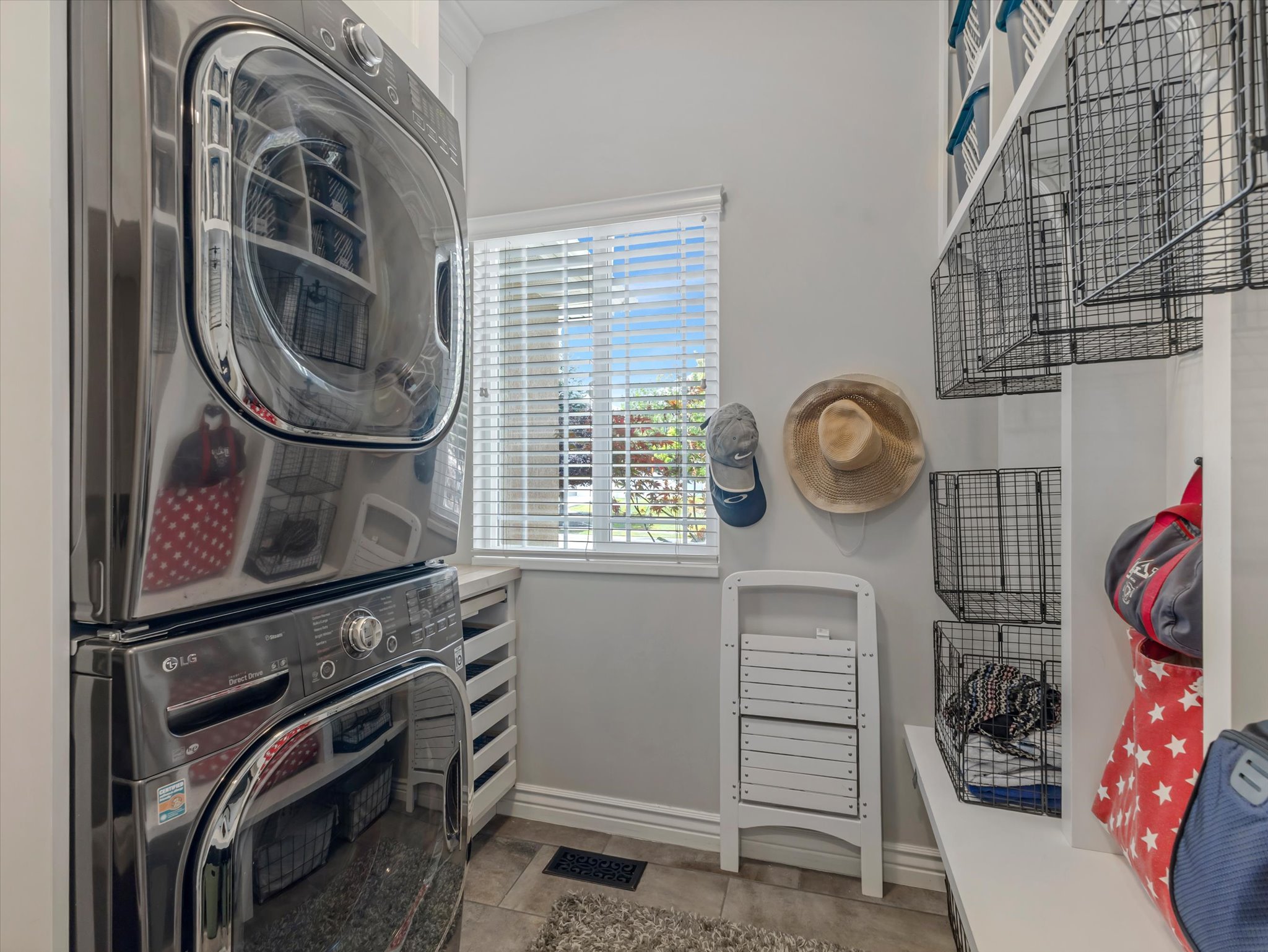 Main floor laundry area featuring stacked appliances, a bright window with blinds, tile flooring, built-in open shelving, and integrated wire baskets for storage