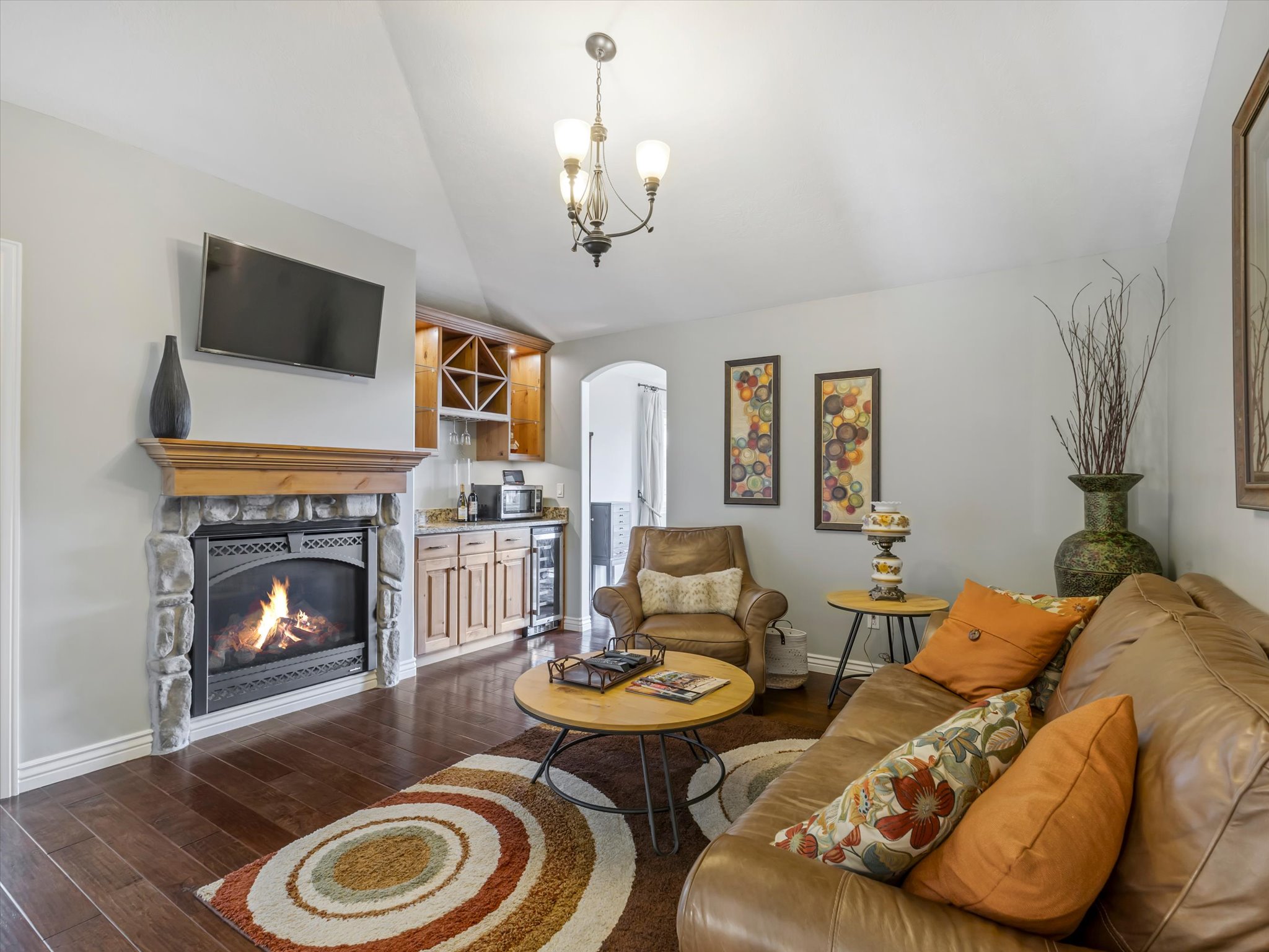 Primary suite living area featuring a stone-surround fireplace with a wood mantle, rich wood-finish flooring, a built-in wet bar with cabinetry and wine storage, and a vaulted ceiling with a chandelier