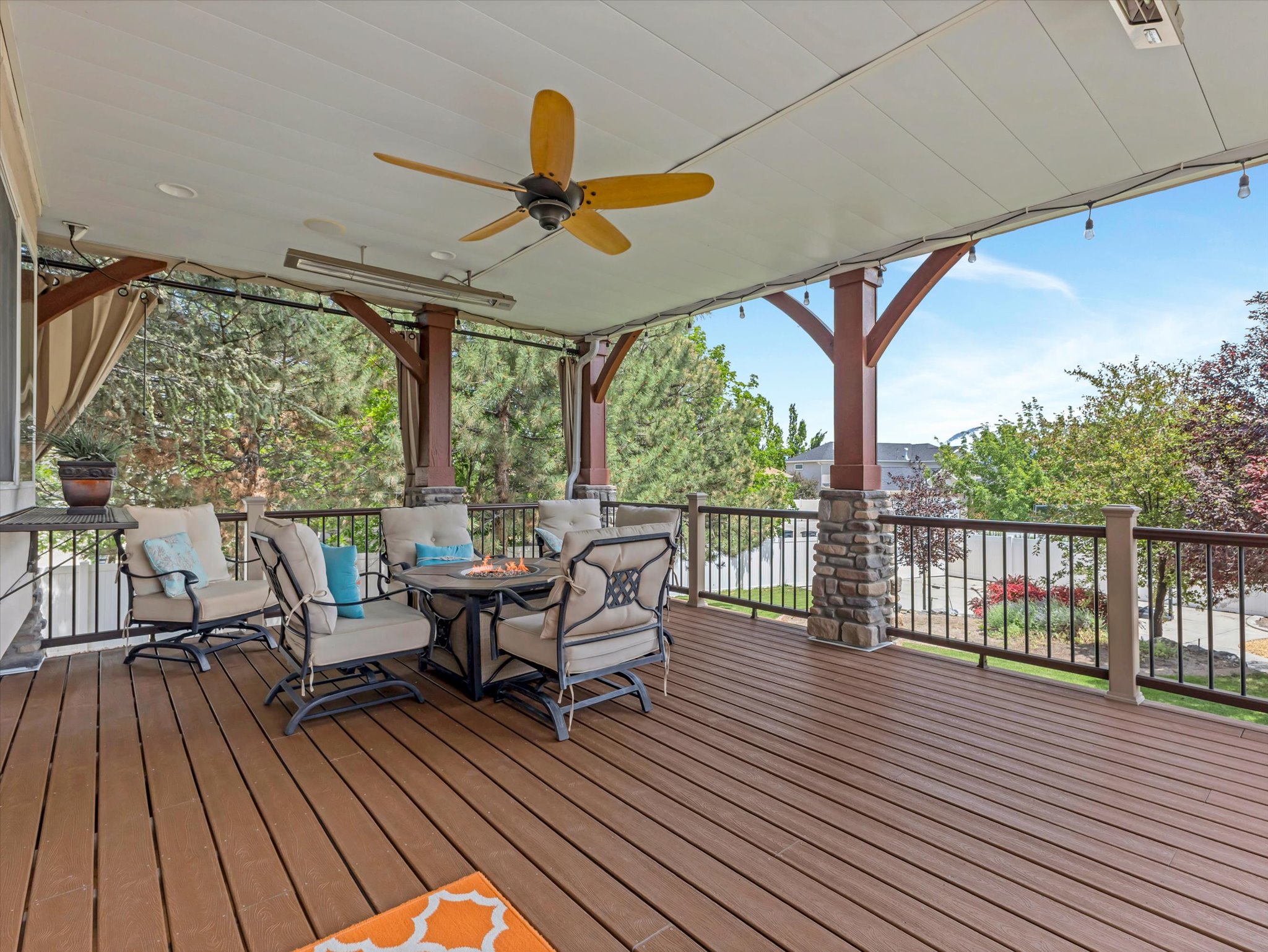 Just off the kitchen, a spacious main floor covered deck featuring composite wood-finish decking, decorative stone-wrapped pillars, a ceiling fan, recessed lighting, and built-in outdoor heaters