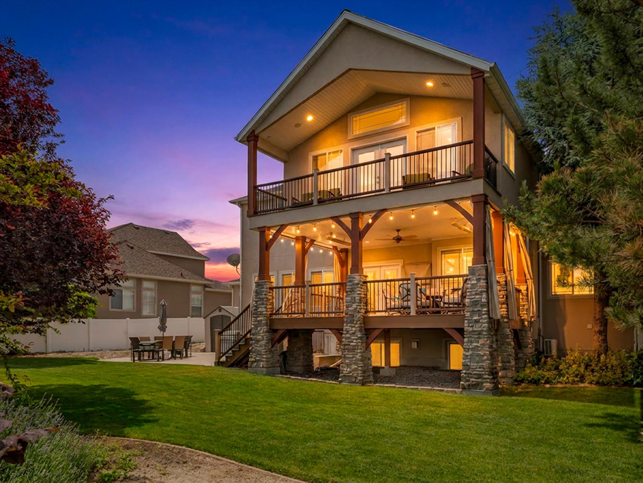 Backyard exterior featuring stone-clad pillars, a lower covered deck with outdoor lights, and the upper, primary suite open-air balcony