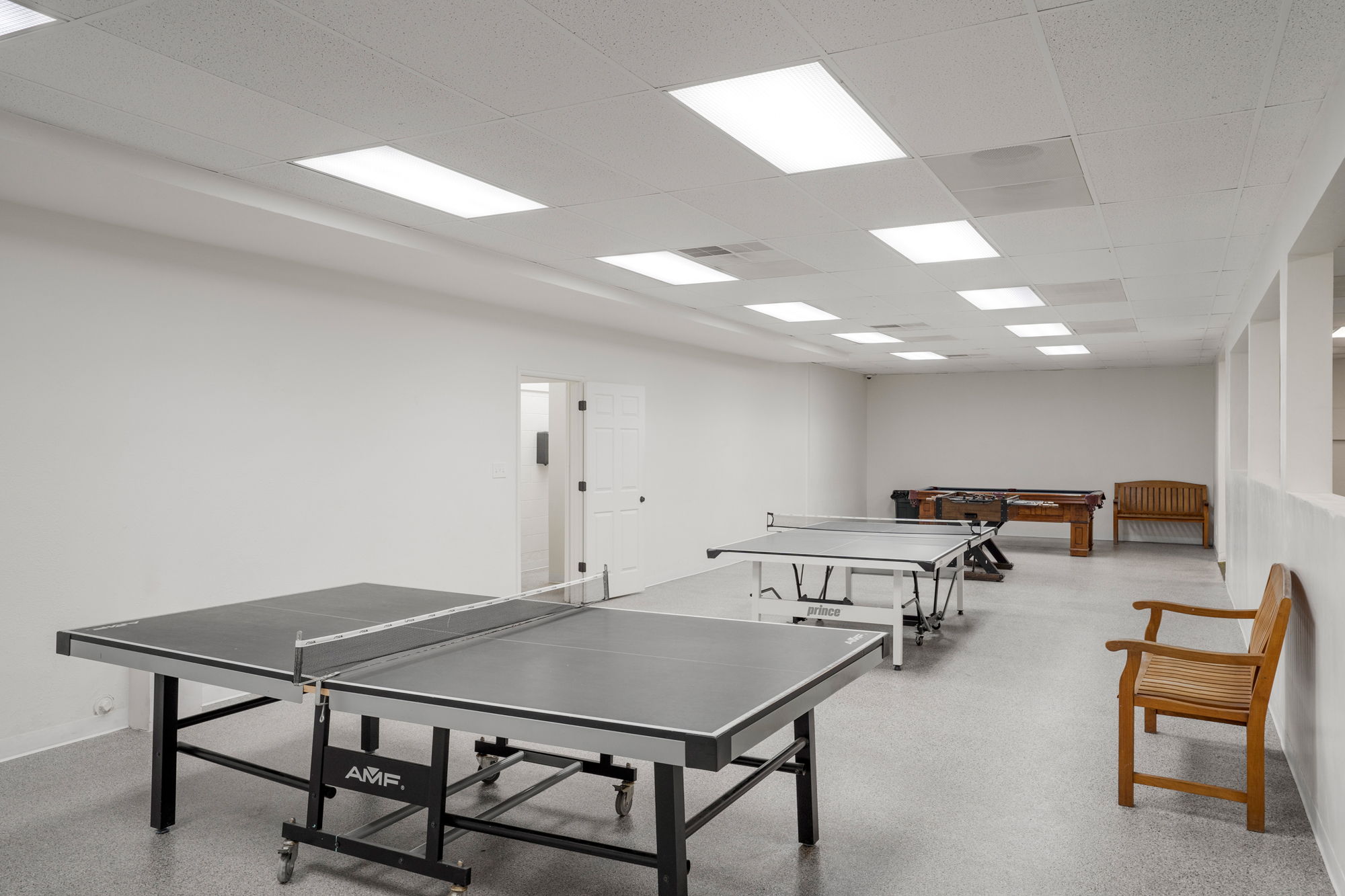 Spacious recreational area featuring light gray speckled flooring, white walls, and a white acoustical tile ceiling with multiple recessed rectangular light fixtures