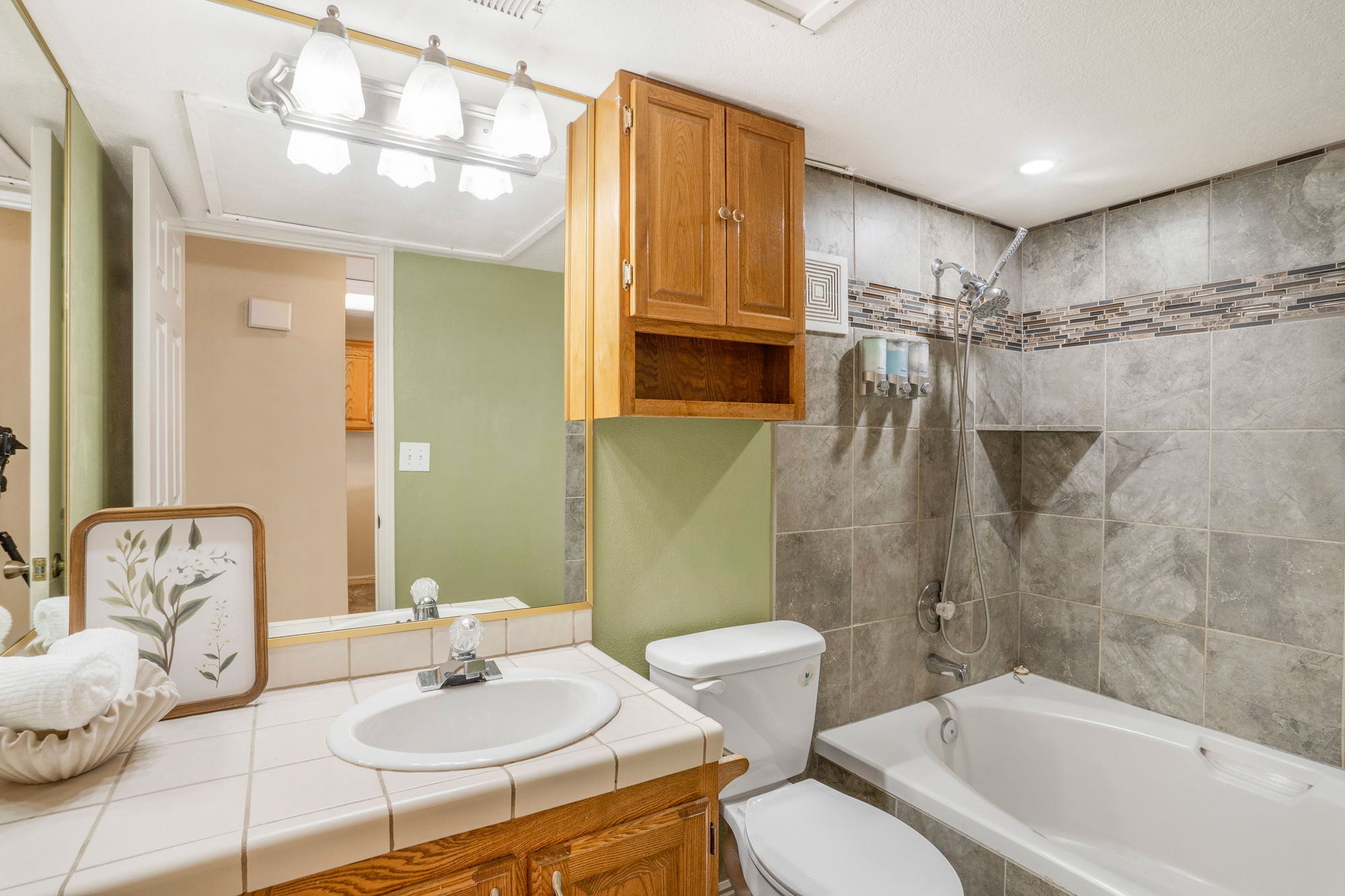 Bathroom featuring a tiled vanity with integrated sink, wood-finish cabinetry, a full-width mirror, and a combination shower-tub with tiled surround and decorative accent band