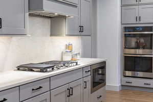 Modern kitchen featuring light gray cabinetry with matte black hardware, white countertops, a built-in gas cooktop, a stainless steel range hood, and a built-in double oven