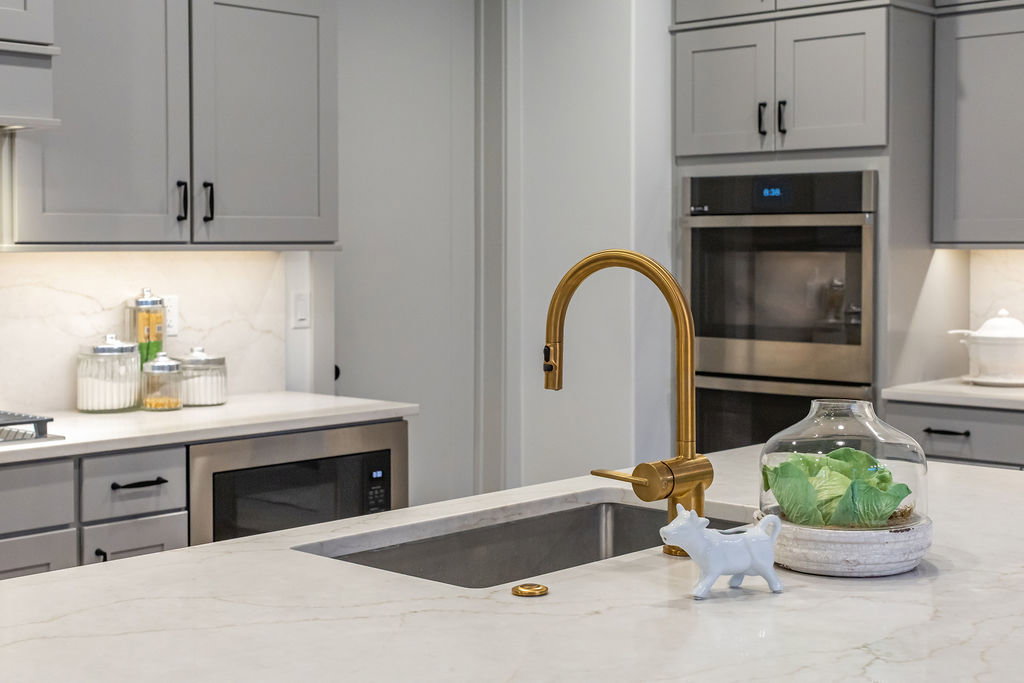 Kitchen featuring light gray cabinetry with dark hardware, a brass-finish gooseneck faucet, a double wall oven, a built-in microwave, and light-toned countertops with an integrated sink