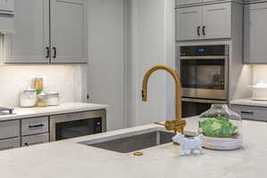 Kitchen featuring light gray cabinetry with dark hardware, a brass-finish gooseneck faucet, a double wall oven, a built-in microwave, and light-toned countertops with an integrated sink