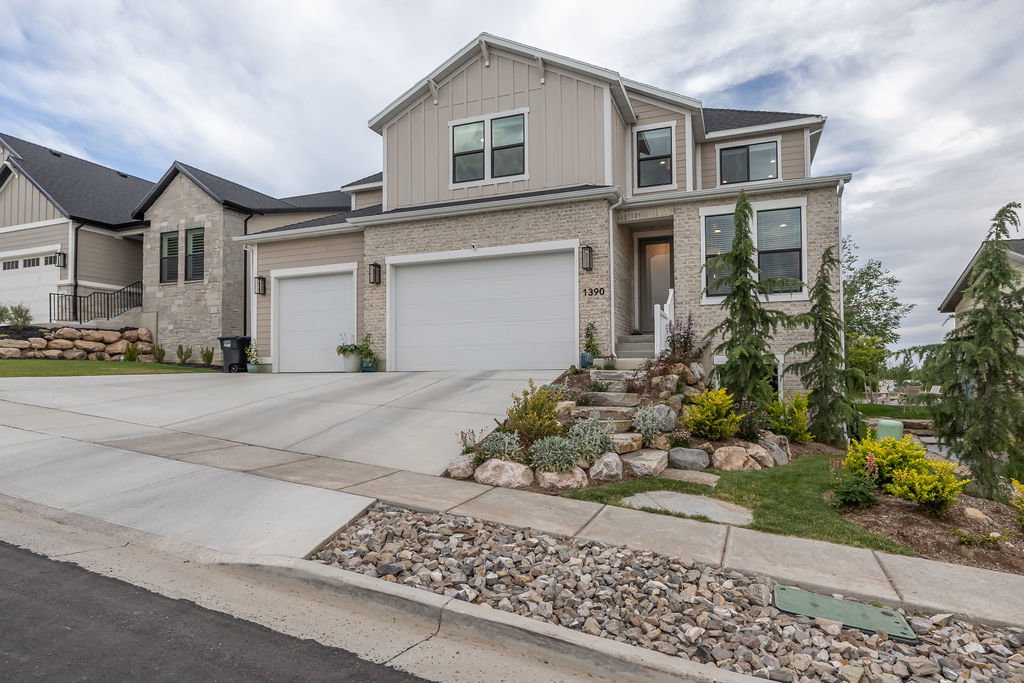 Modern residential exterior featuring stone and board-and-batten siding, a two-car garage, a concrete driveway, and a landscaped rock garden with established plantings