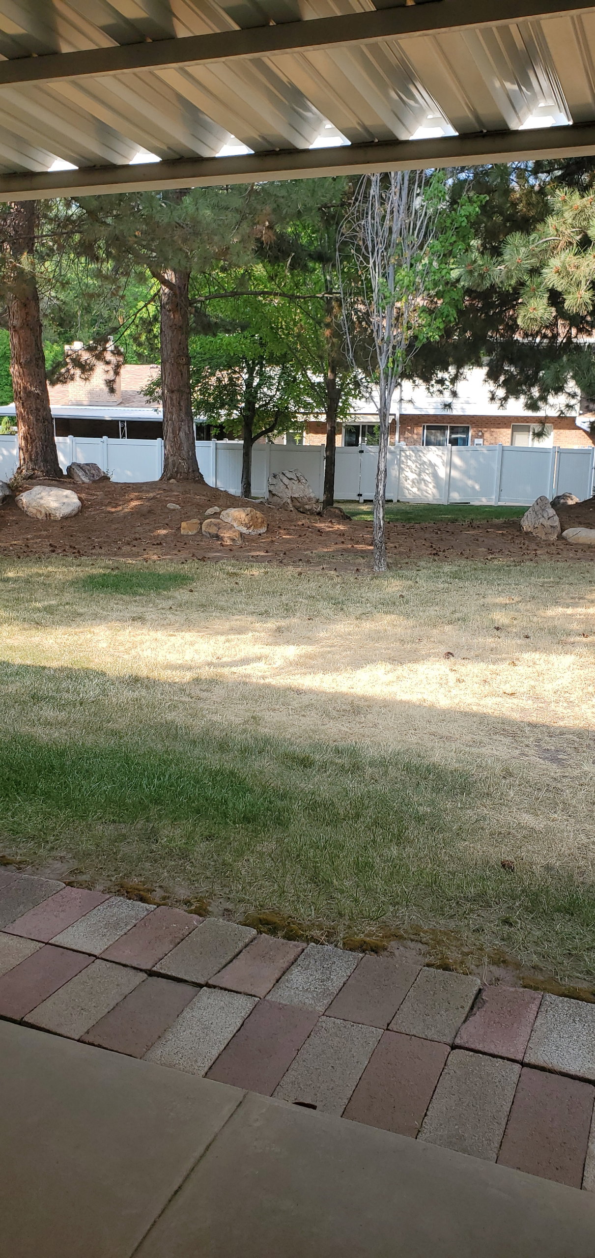 Covered patio featuring a corrugated metal roof and brick paver border, overlooking a lawn area with mature trees and a white privacy fence