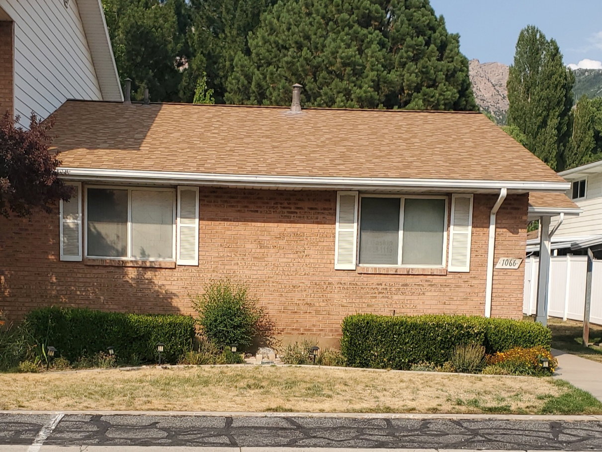 Brick exterior with multiple windows featuring louvered shutters, a shingle roof, and established front landscaping