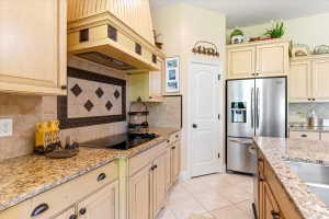 Kitchen featuring light-toned cabinetry with dark hardware, granite countertops, a tiled backsplash with decorative accents, an electric cooktop, and a stainless steel refrigerator