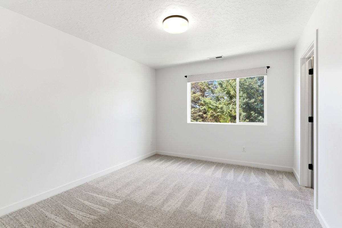 Bright interior room featuring a window with roller shade, white trim, light-toned patterned carpet, flush-mount ceiling lighting, and a white interior door with black hardware
