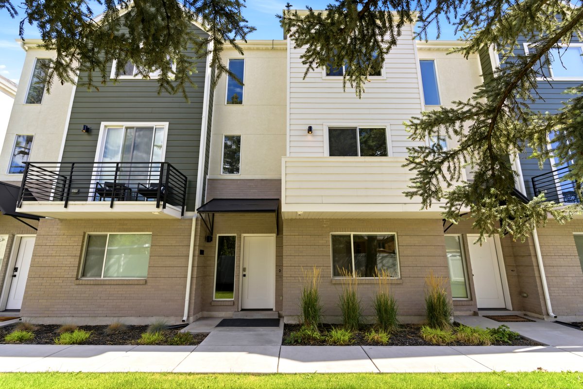 Modern townhome exterior featuring an upper-level balcony with black metal railings, a contemporary awning over the entrance, and a light-colored brick facade on the lower level