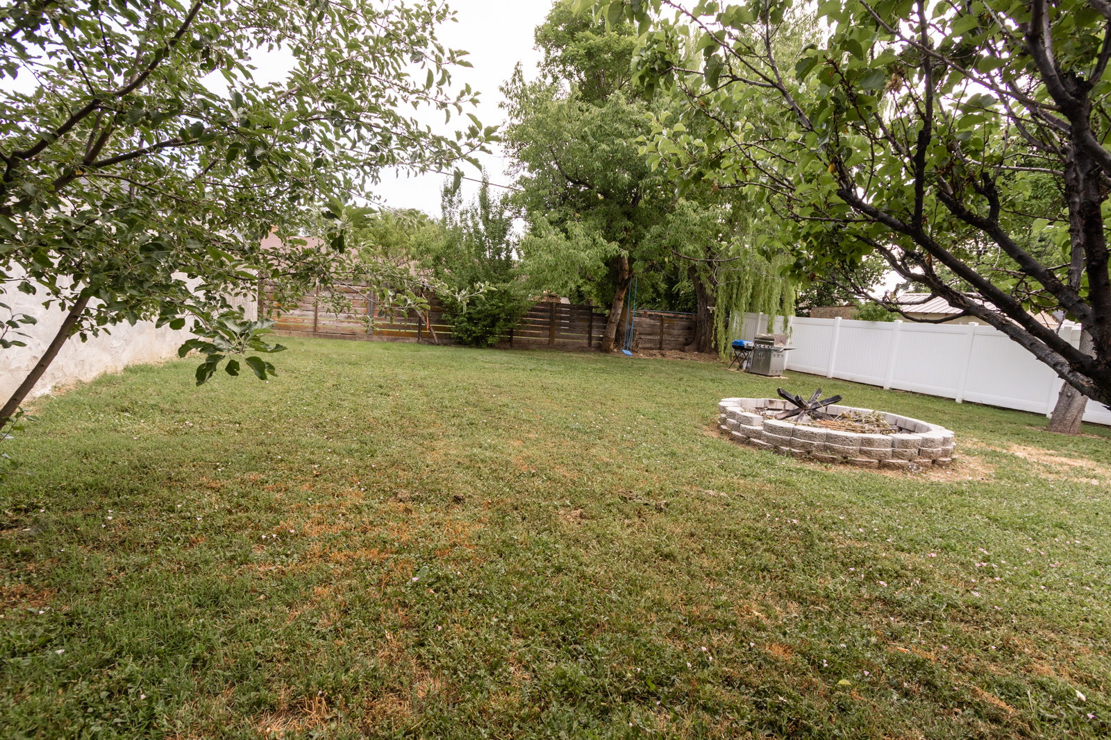 Expansive backyard featuring a stone-lined fire pit, mature trees, and a combination of wood and white vinyl fencing