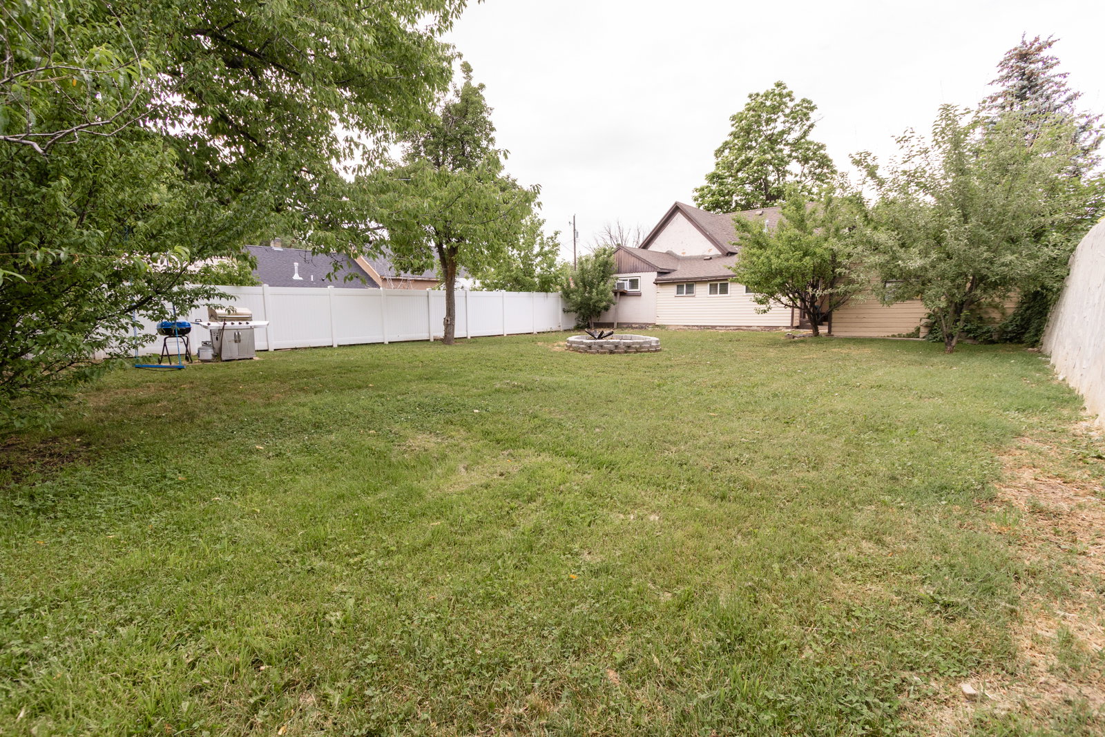 Expansive backyard featuring a manicured lawn, established trees, a white vinyl privacy fence, and a stone-lined fire pit