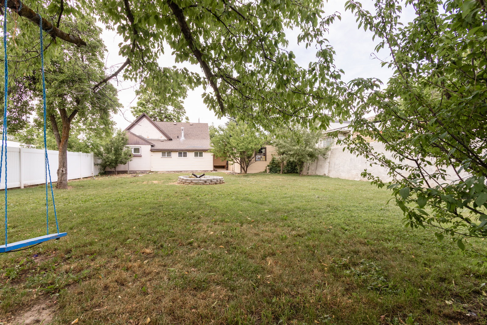 Expansive backyard featuring a stone-ringed fire pit, mature trees, and a white privacy fence