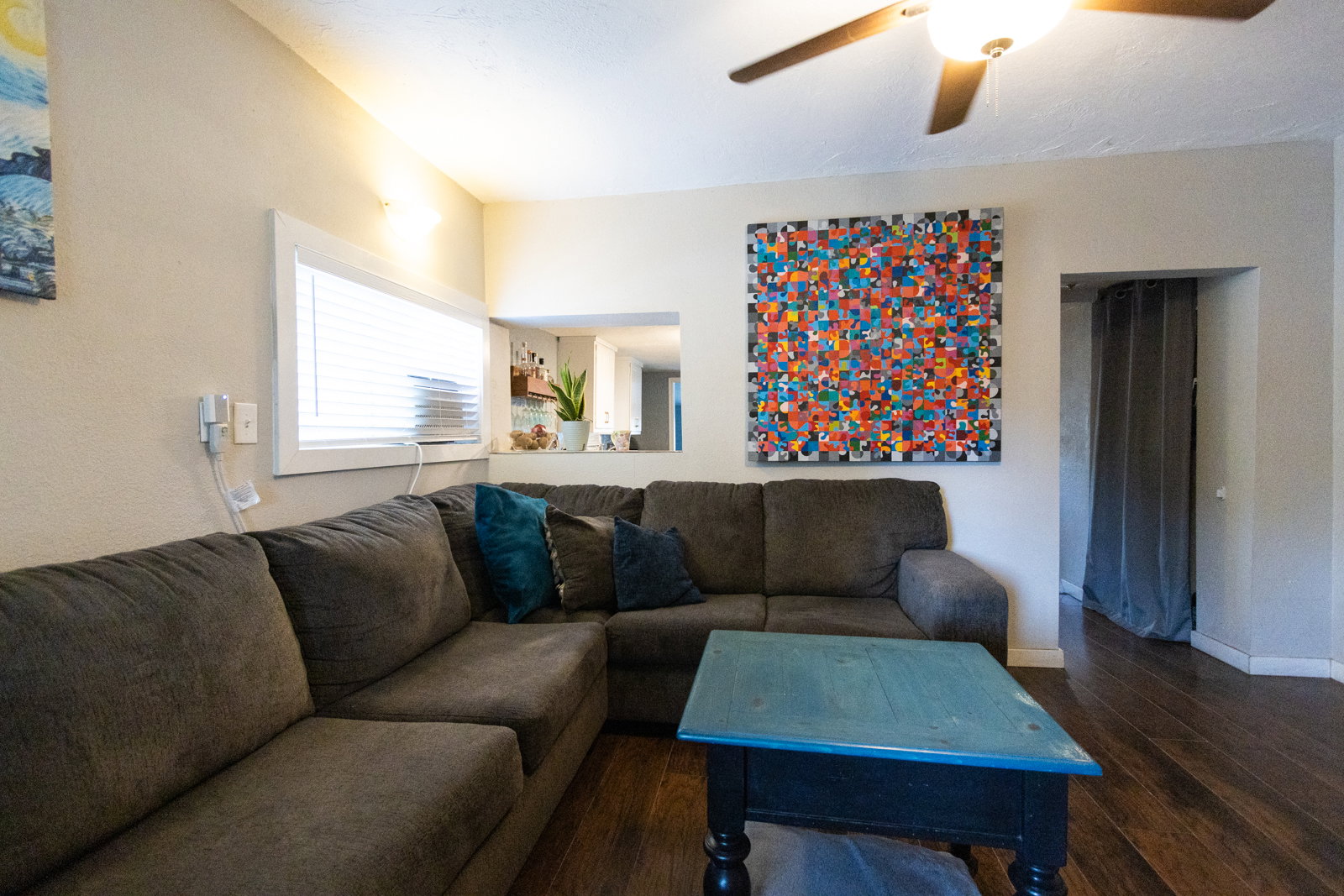 Living area featuring wood-finish flooring, a white-framed window with blinds, an overhead ceiling fan with lighting, and a pass-through opening to another room