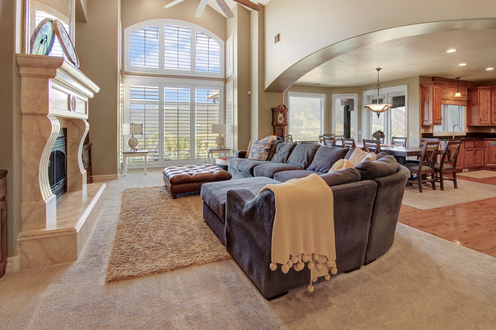 Living area featuring a floor-to-ceiling window array with plantation shutters, a fireplace with a stone mantel, vaulted ceilings, and an arched entry to a dining space with wood-finish flooring