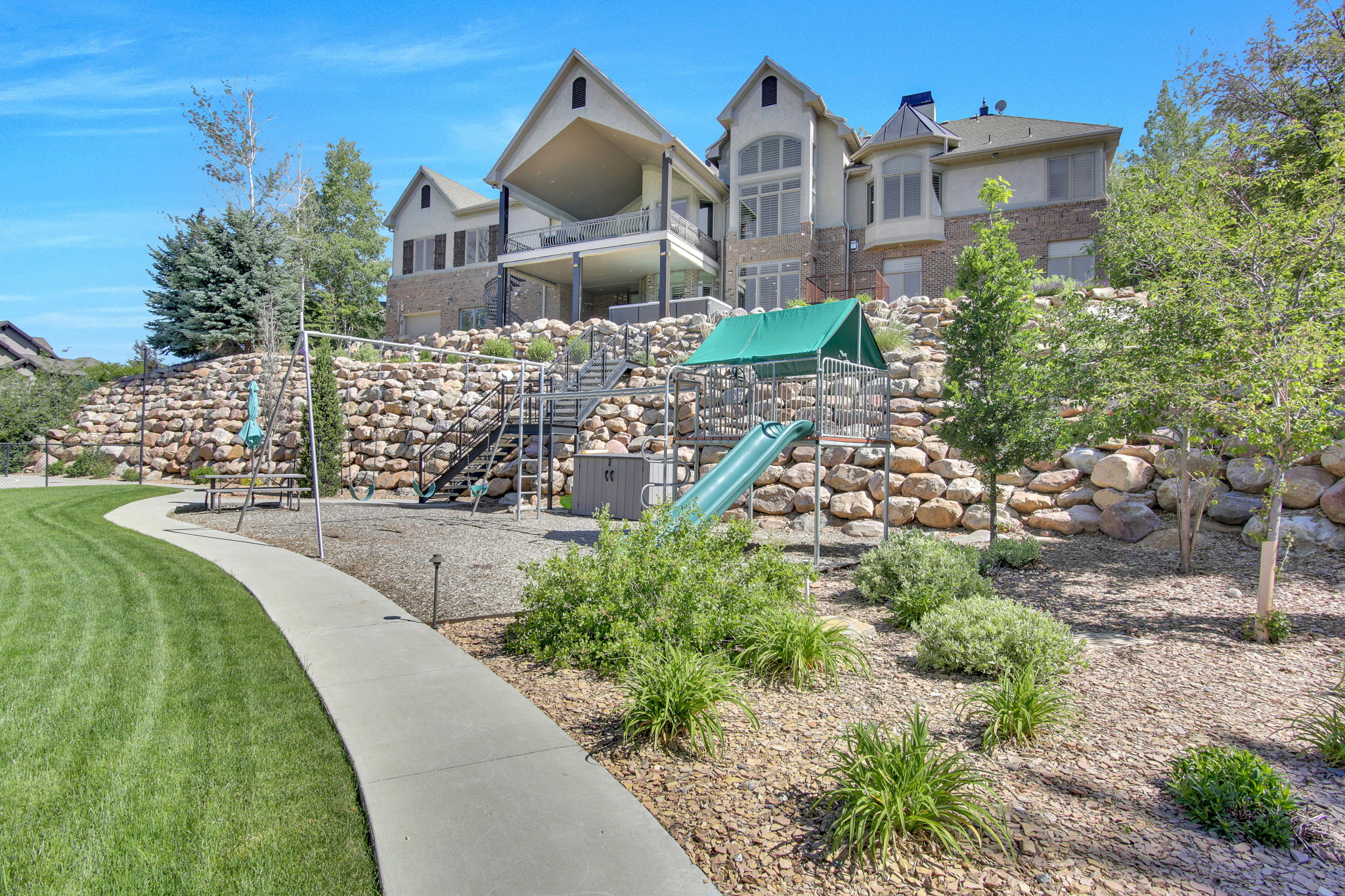 Expansive terraced backyard featuring a manicured lawn, concrete pathway, rock retaining walls, a multi-level deck, and a dedicated play structure