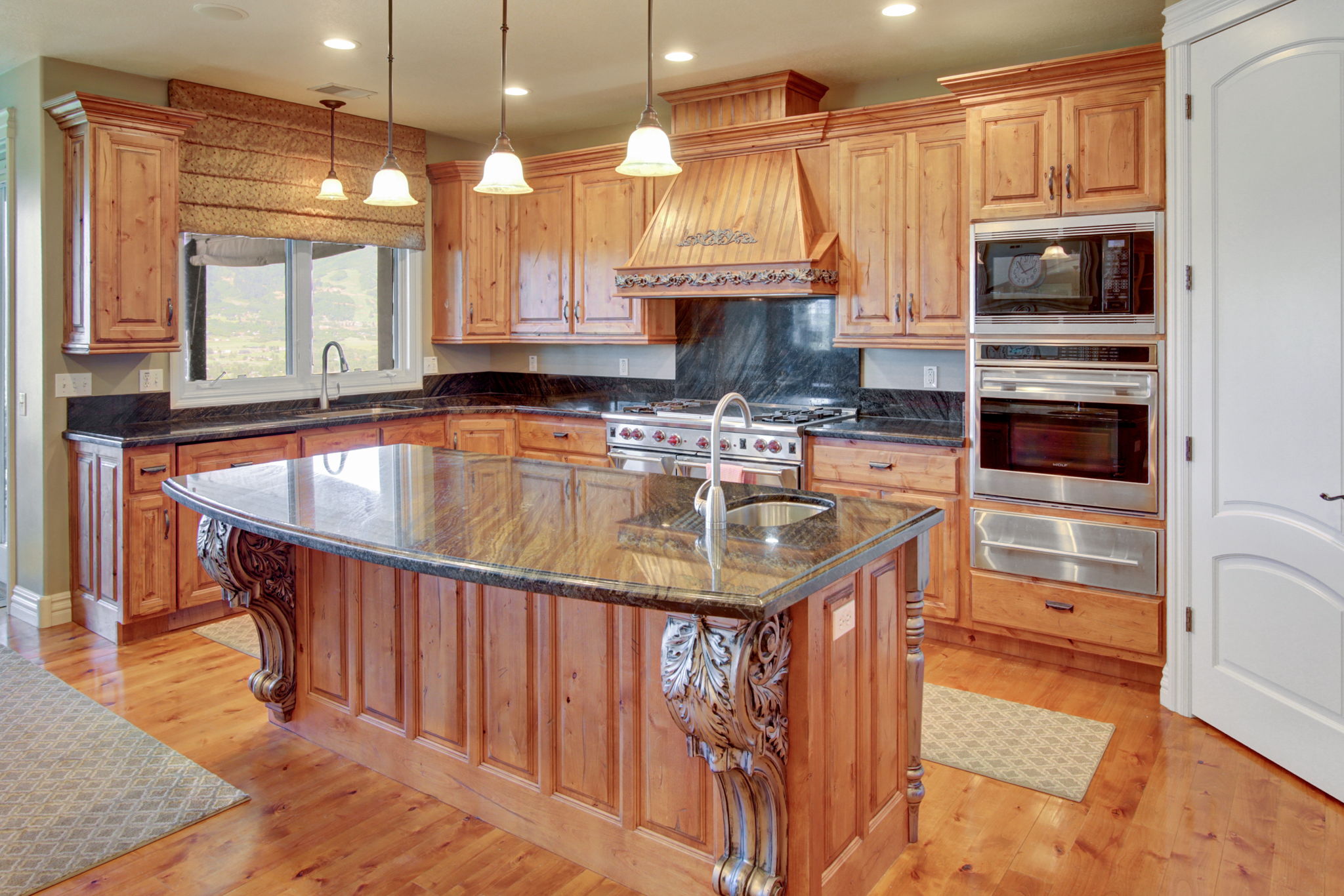 Expansive kitchen featuring a large central island with an integrated sink, rich wood-finish cabinetry, dark stone countertops, stainless steel appliances, and wood-finish flooring