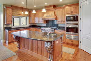 Expansive kitchen featuring a large central island with an integrated sink, rich wood-finish cabinetry, dark stone countertops, stainless steel appliances, and wood-finish flooring