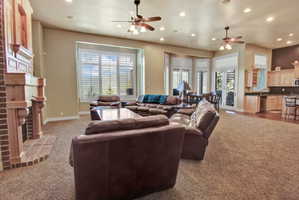Spacious open-concept living area featuring a brick fireplace with a wood mantel, large windows with white louvered shutters, recessed lighting, and ceiling fans