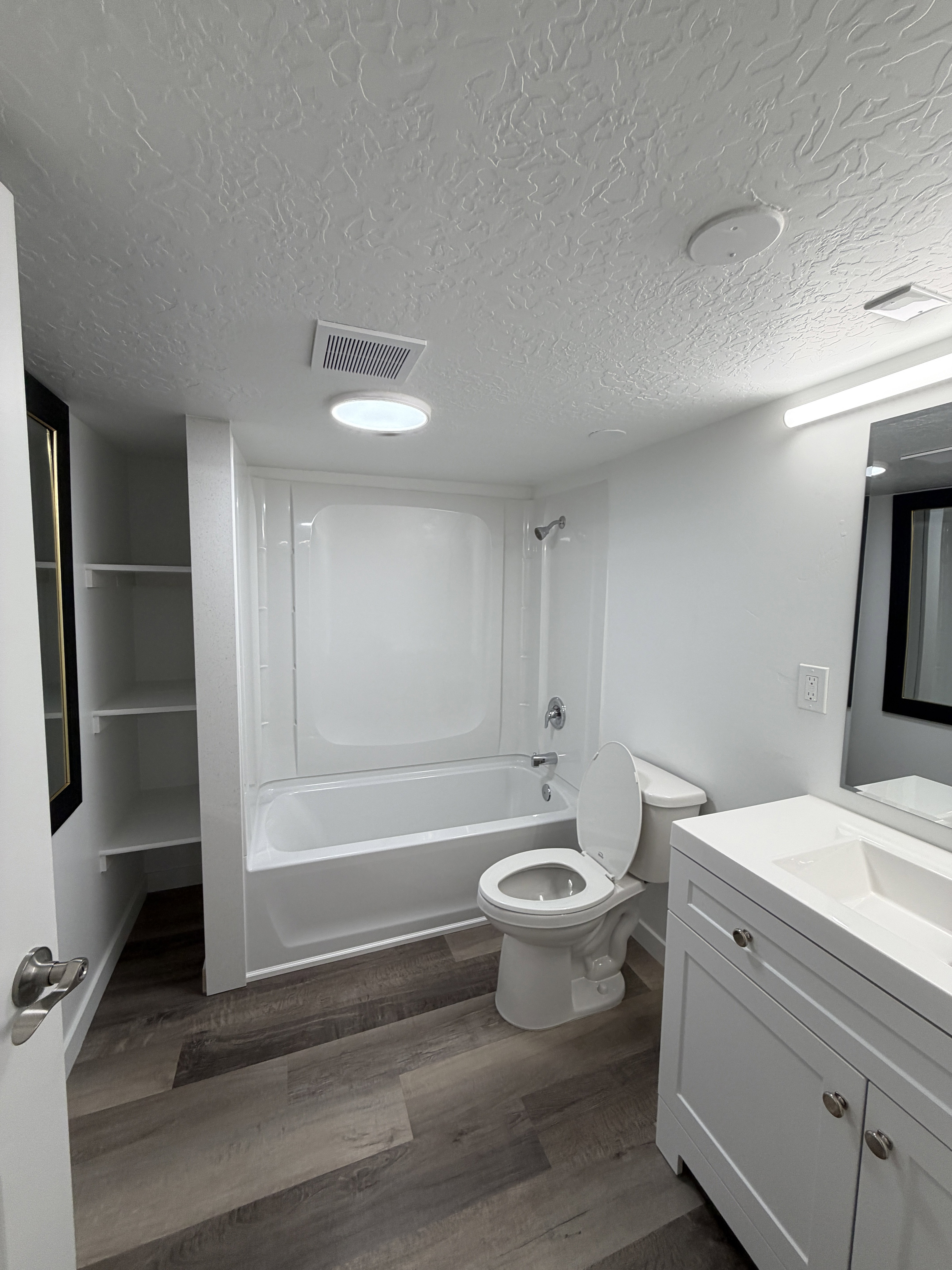 Bathroom featuring wood-finish flooring, a white vanity with a single basin sink, a white bathtub with a shower surround, and integrated shelving