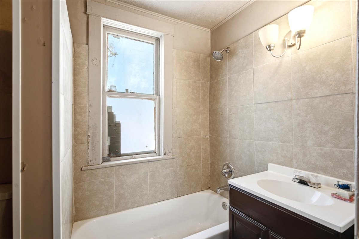 Bathroom featuring a white bathtub, ceramic tile walls, a dark wood-finish vanity with an integrated sink, a chrome faucet, and a wall-mounted light fixture