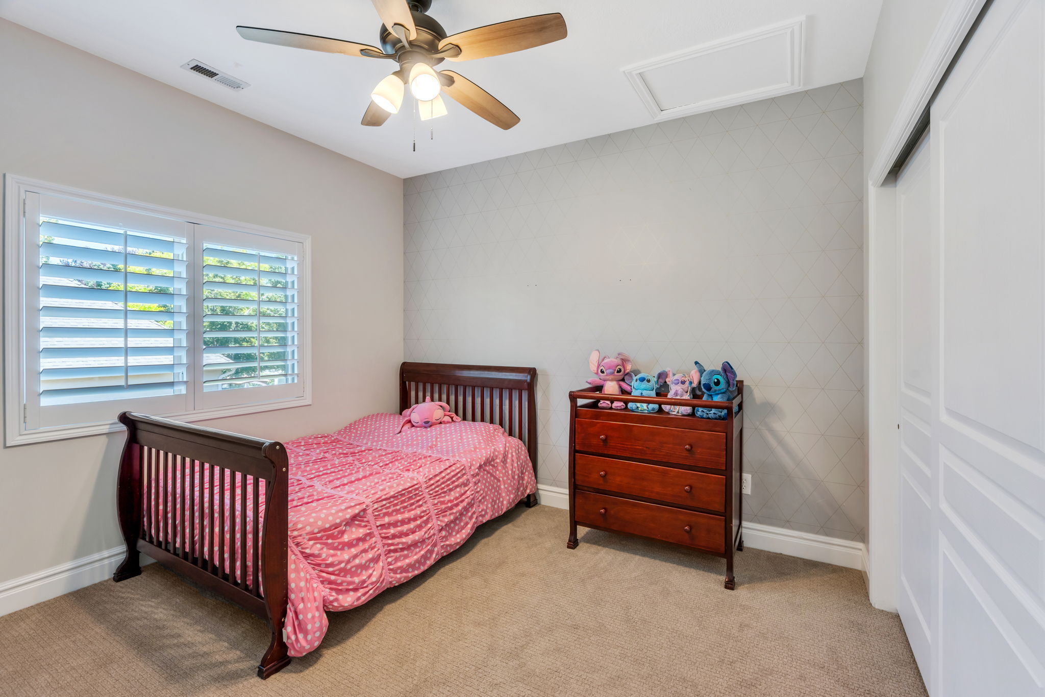 Carpeted room featuring a ceiling fan with light fixture, window with white plantation shutters, patterned accent wall, and sliding closet doors