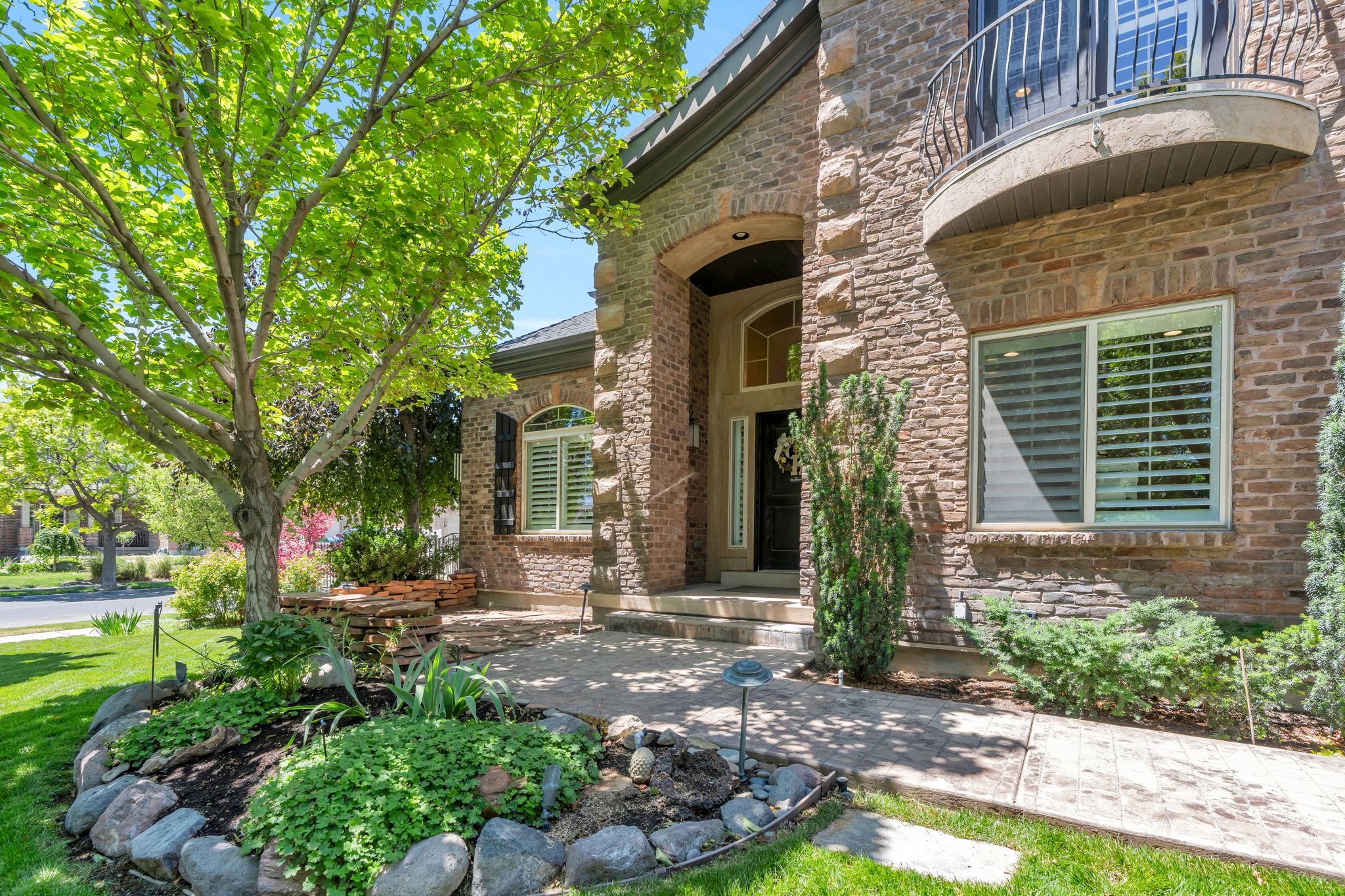 Brick and stone facade residence featuring an arched entryway, decorative balcony, and a manicured front yard with mature trees