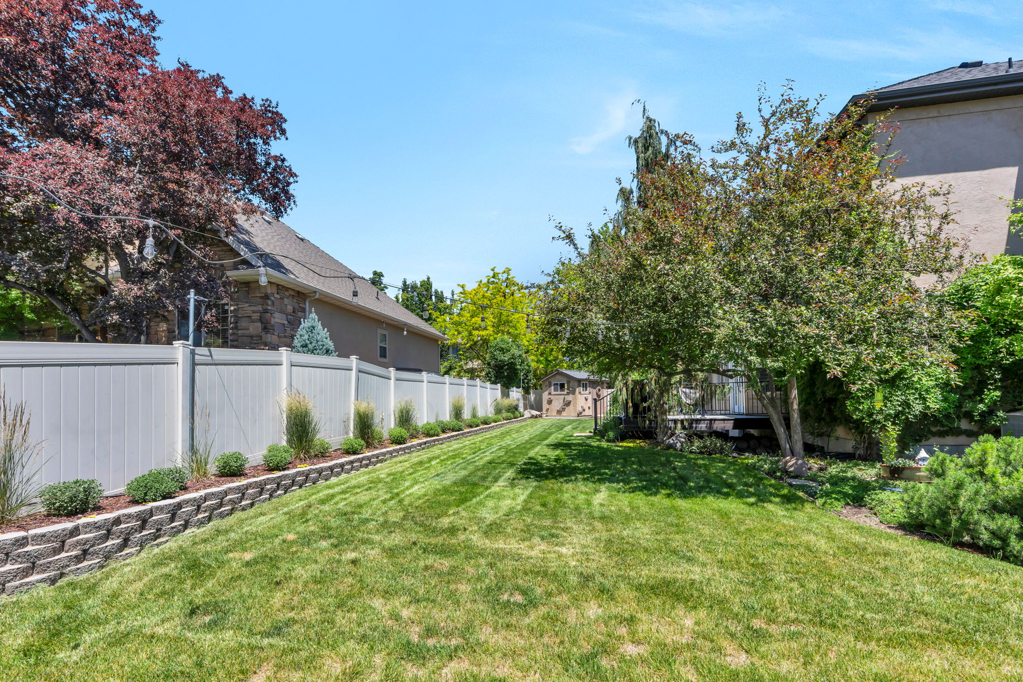 Expansive backyard featuring a manicured lawn, a retaining wall with structured landscaping, a white privacy fence, mature trees, and an outdoor storage shed
