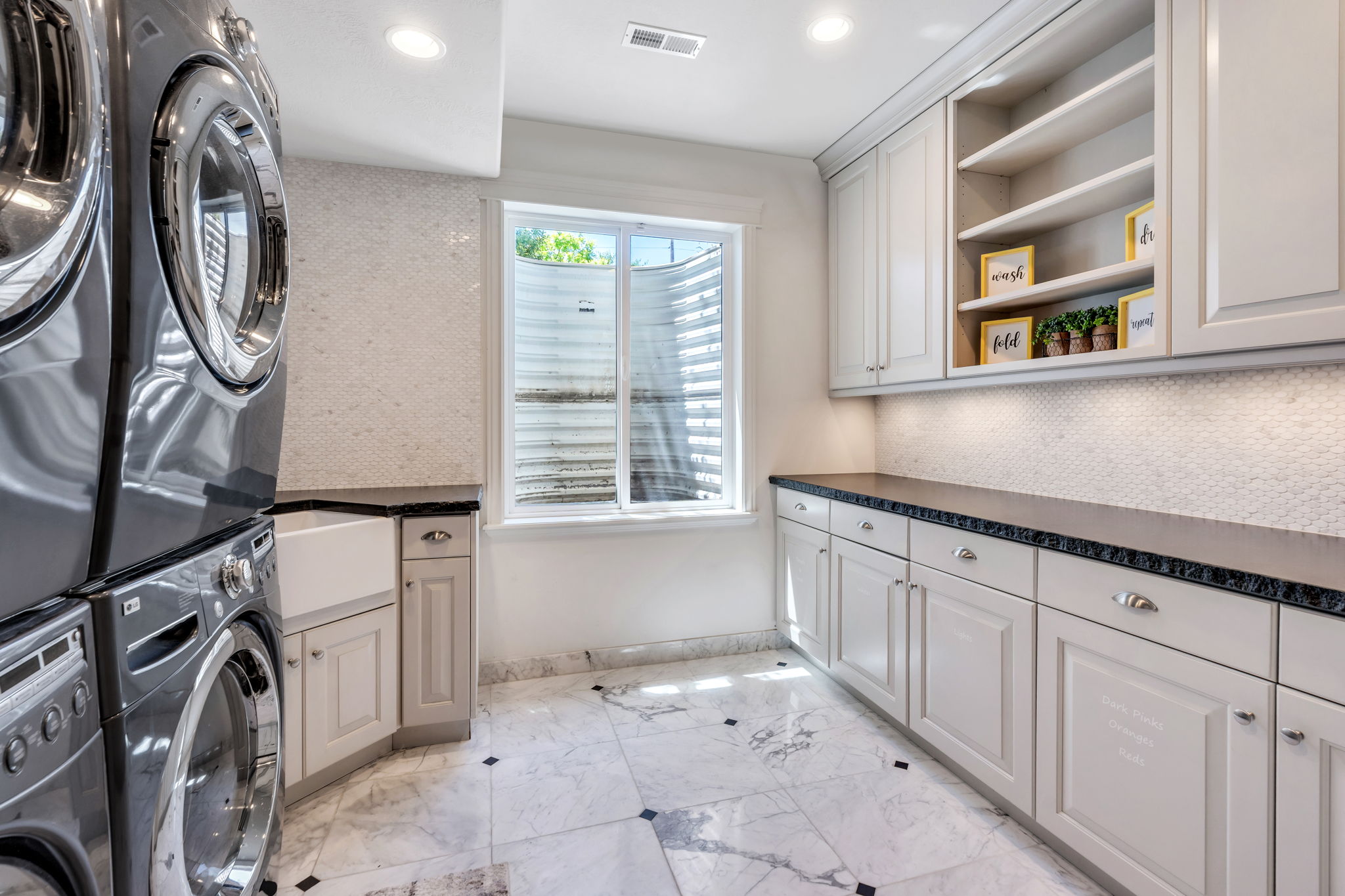 Spacious laundry area featuring stacked appliances, a large farmhouse sink, extensive built-in cabinetry, dark countertops, and marble-look tile flooring with black square inlays