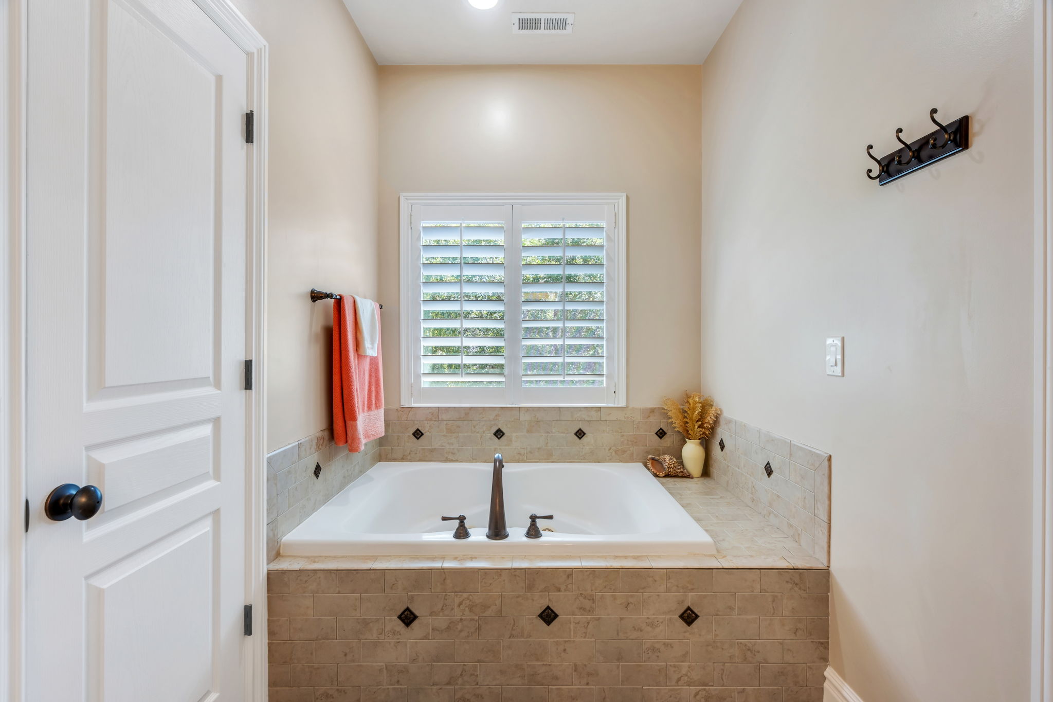 Built-in soaking tub with a tiled surround featuring dark accent squares, a window with plantation shutters, and oil-rubbed bronze fixtures