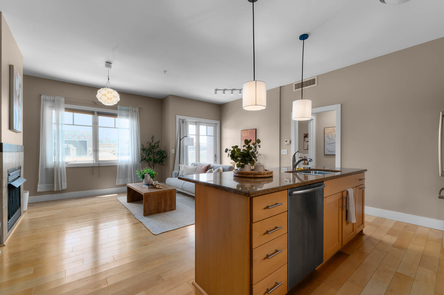 Open-concept living space featuring wood-finish flooring, a kitchen island with integrated sink and dishwasher, pendant lighting, a fireplace with dark tile surround, and multiple windows with white trim