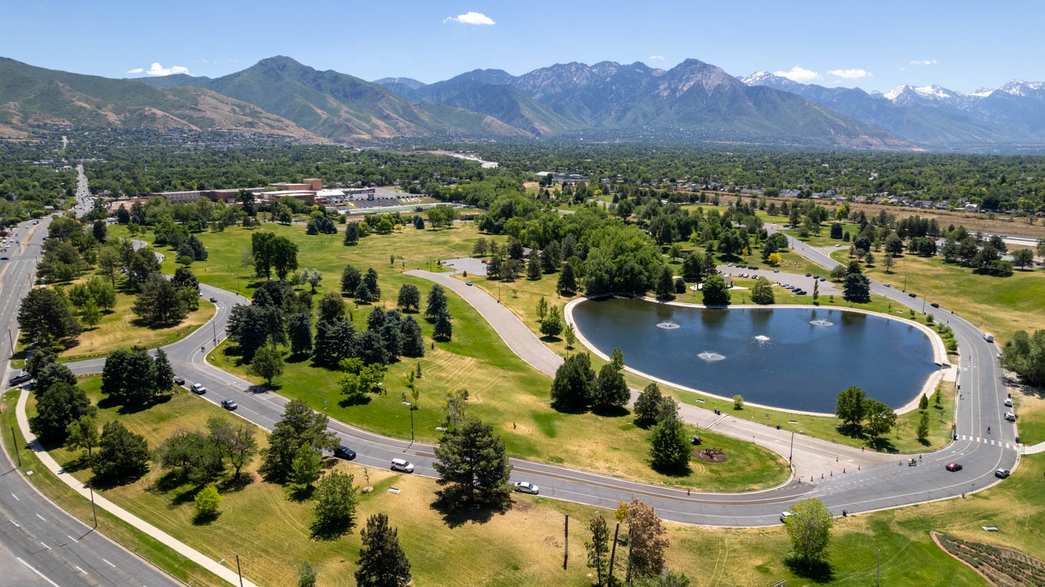 Expansive park grounds featuring a large pond with multiple fountains, paved pathways, mature trees, and mountainous scenery