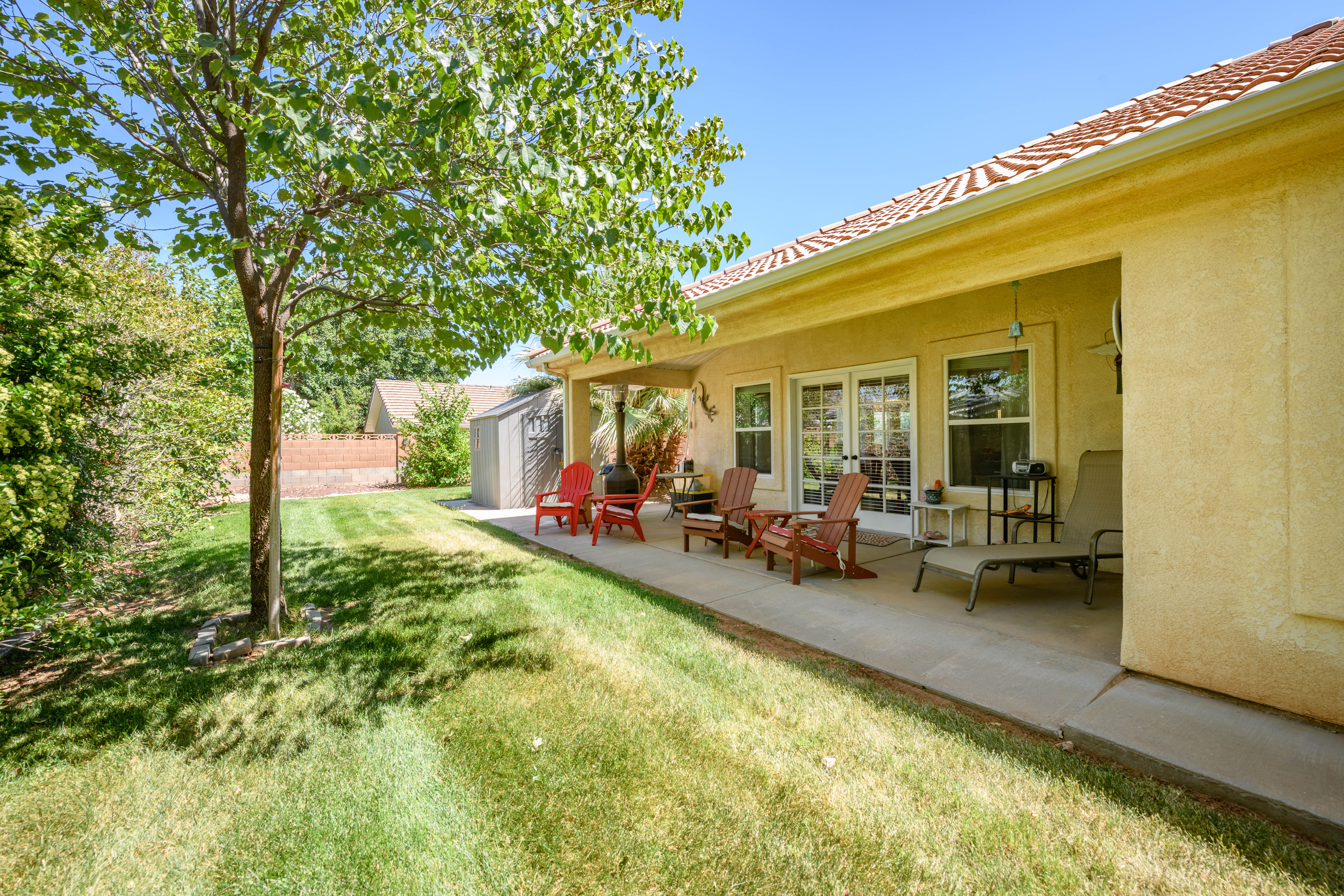 Expansive backyard featuring a covered patio with concrete flooring, stucco exterior, multiple windows, and a mature tree