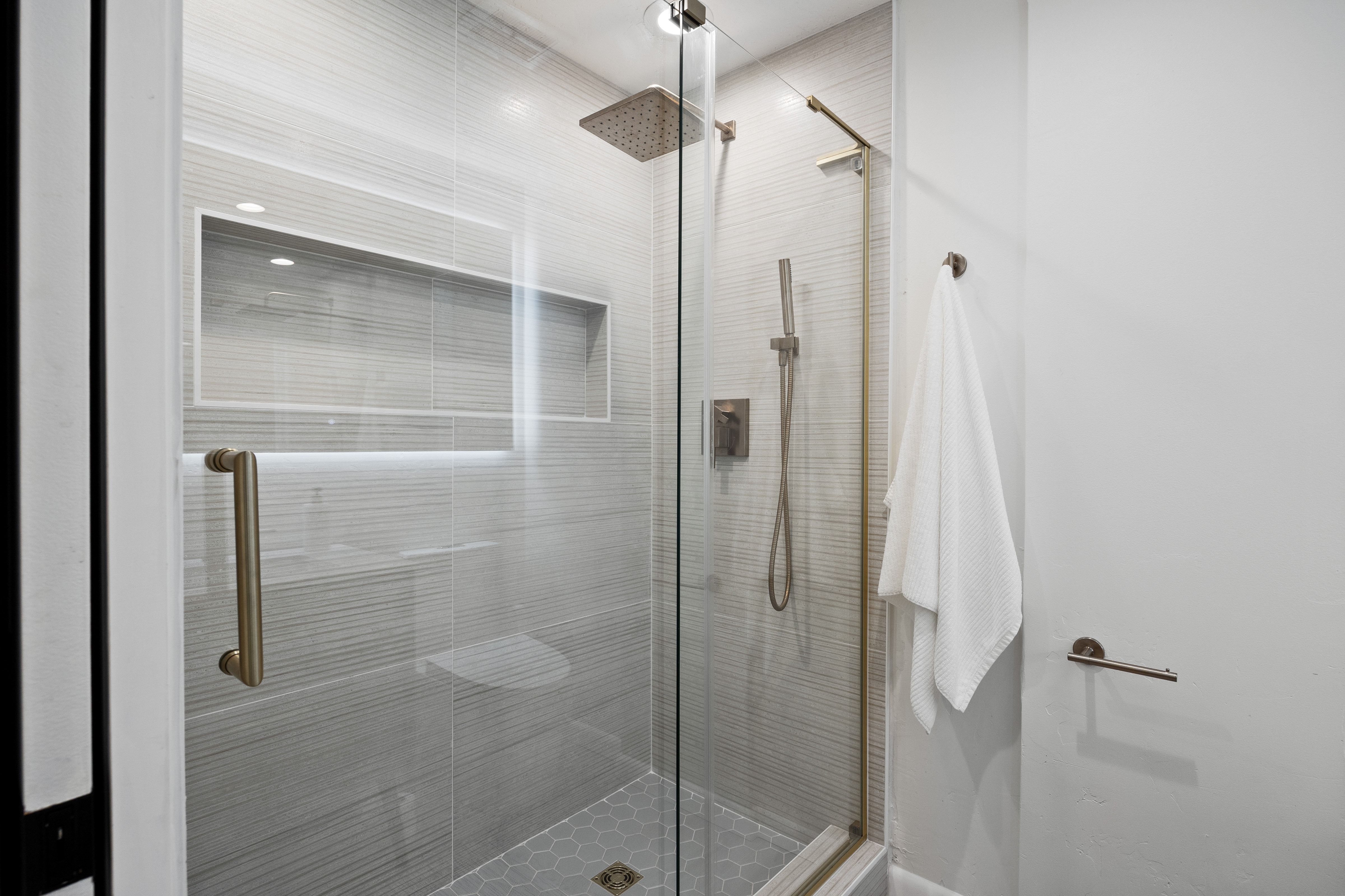 Walk-in shower featuring a frameless glass enclosure, a built-in shower niche with accent lighting, a rain showerhead, a hand-held shower fixture, and hexagonal floor tiling