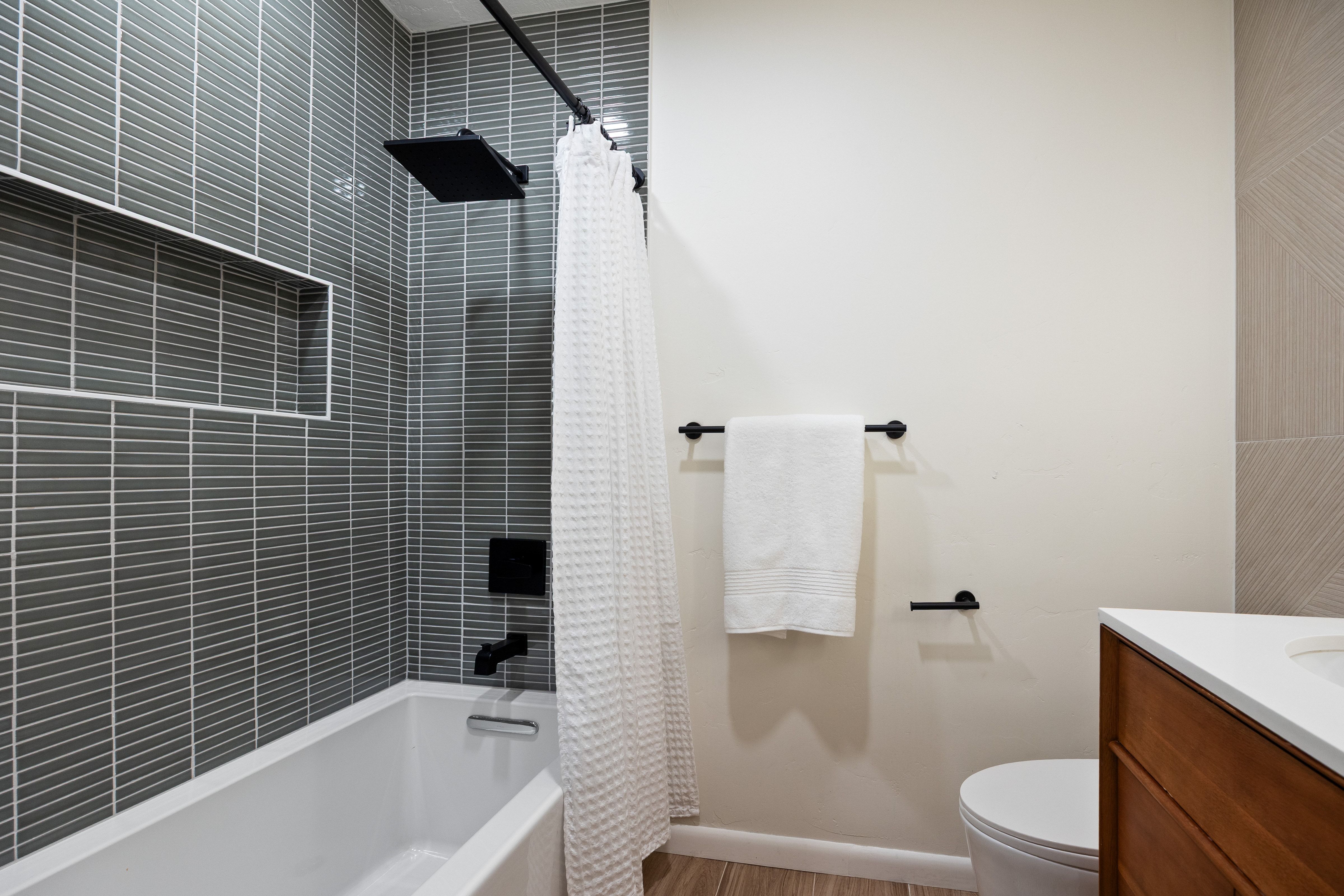 Bathroom featuring a white tub with a matte black rain shower head and fixtures, grey horizontal tile surround with a recessed niche, wood-finish flooring, and a vanity with a white countertop