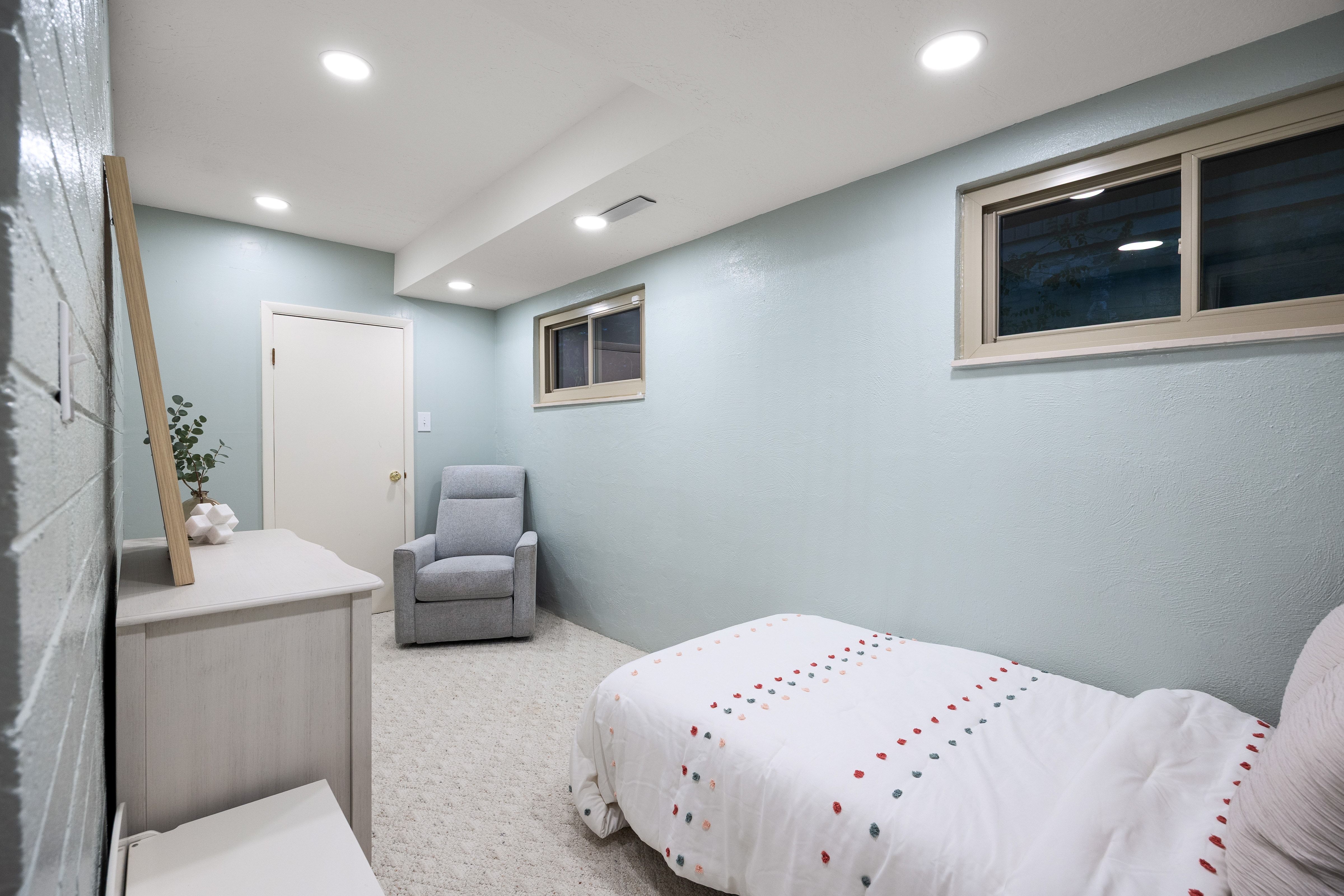 Recessed ceiling lighting illuminates a room featuring light blue walls, a white painted door with brass hardware, and a window with a tan frame
