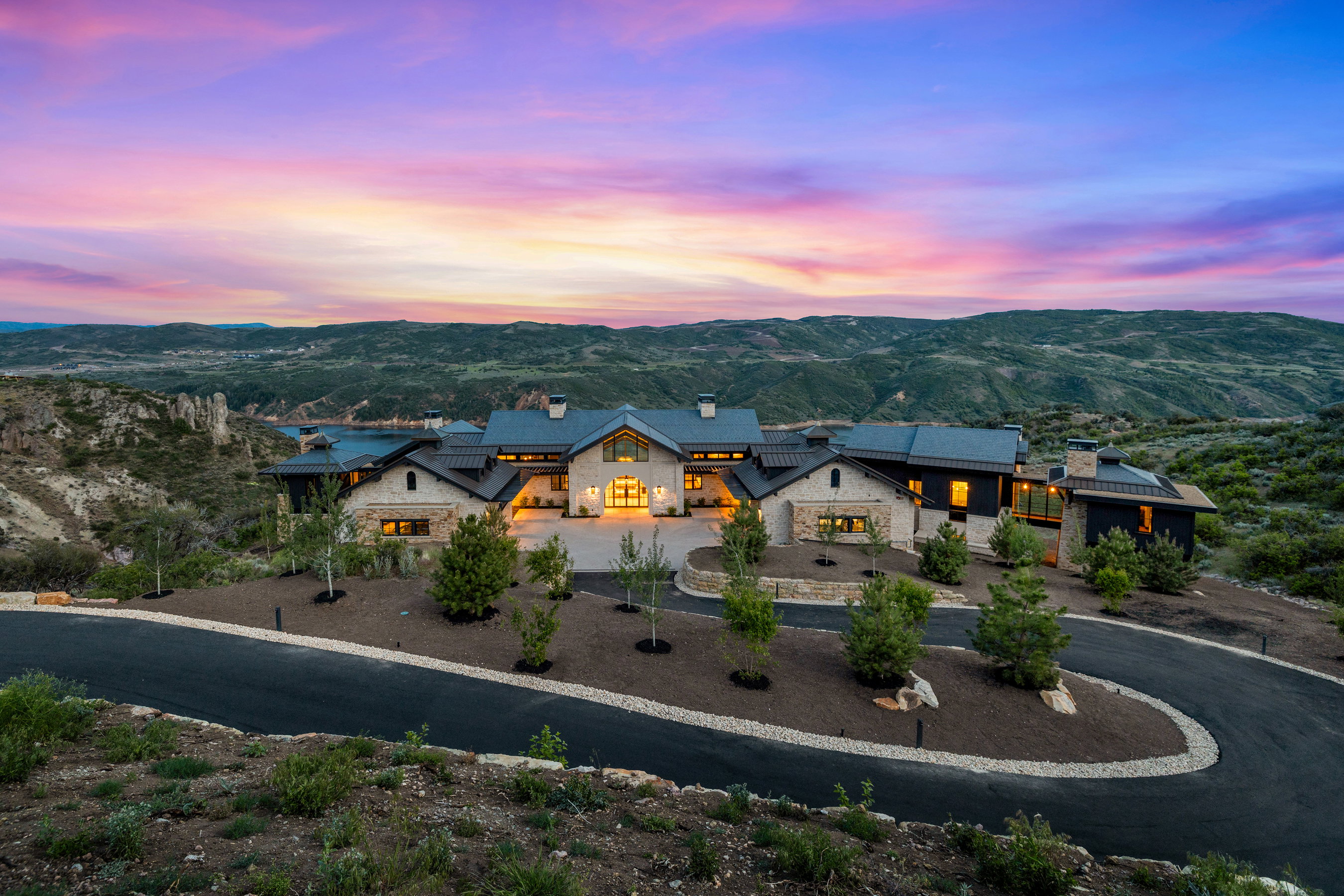 Expansive property featuring a stone and dark-paneled exterior, multiple gabled roofs, a paved circular driveway, and integrated outdoor lighting
