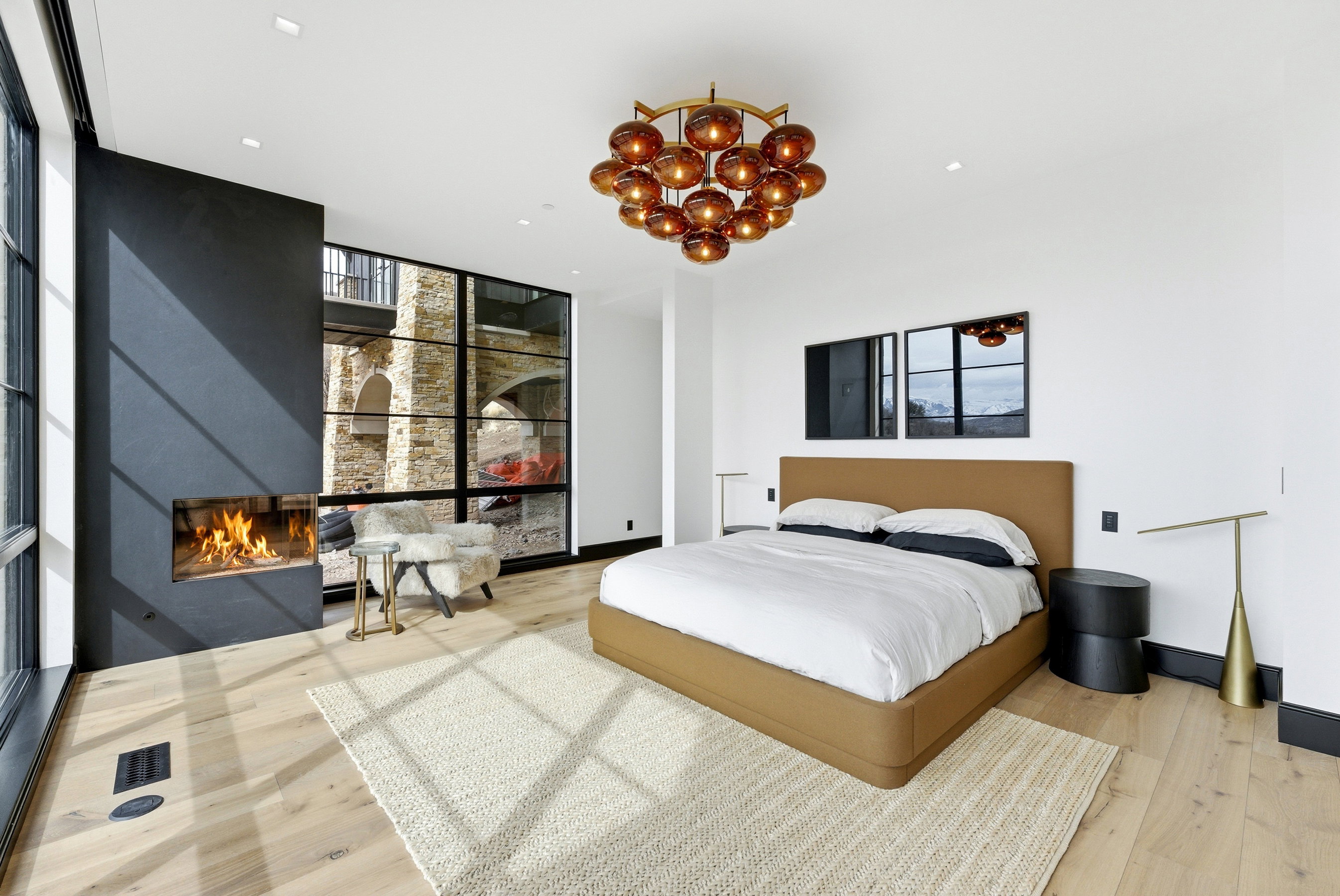 Light wood-finish flooring featuring a modern fireplace with a black surround, large black-framed windows, a distinctive amber-toned chandelier, and recessed lighting