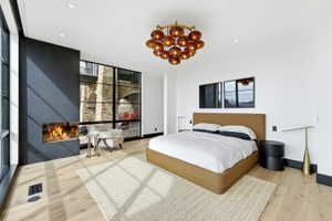 Light wood-finish flooring featuring a modern fireplace with a black surround, large black-framed windows, a distinctive amber-toned chandelier, and recessed lighting