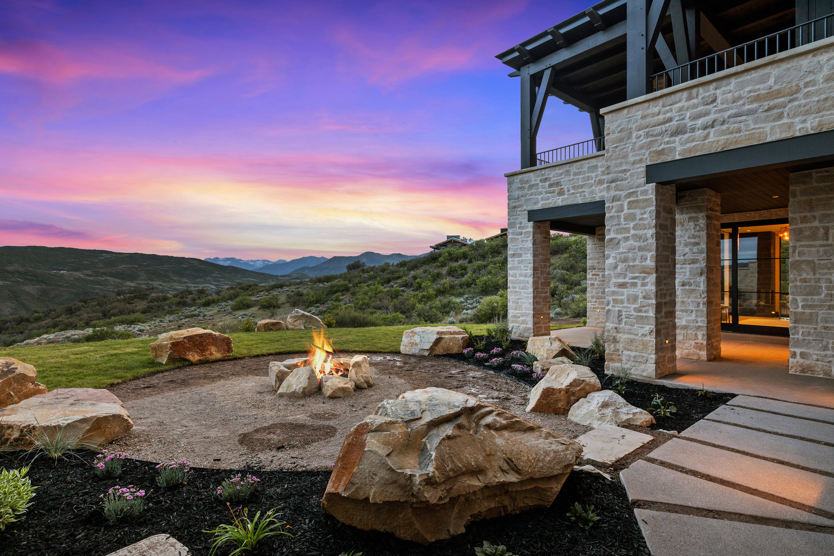 Stone exterior with an upper-level balcony featuring dark wood accents, a ground-level patio with large format pavers, a rock-bordered fire pit, and a manicured lawn