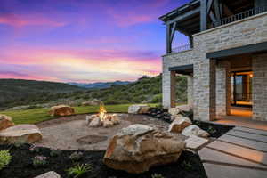 Stone exterior with an upper-level balcony featuring dark wood accents, a ground-level patio with large format pavers, a rock-bordered fire pit, and a manicured lawn