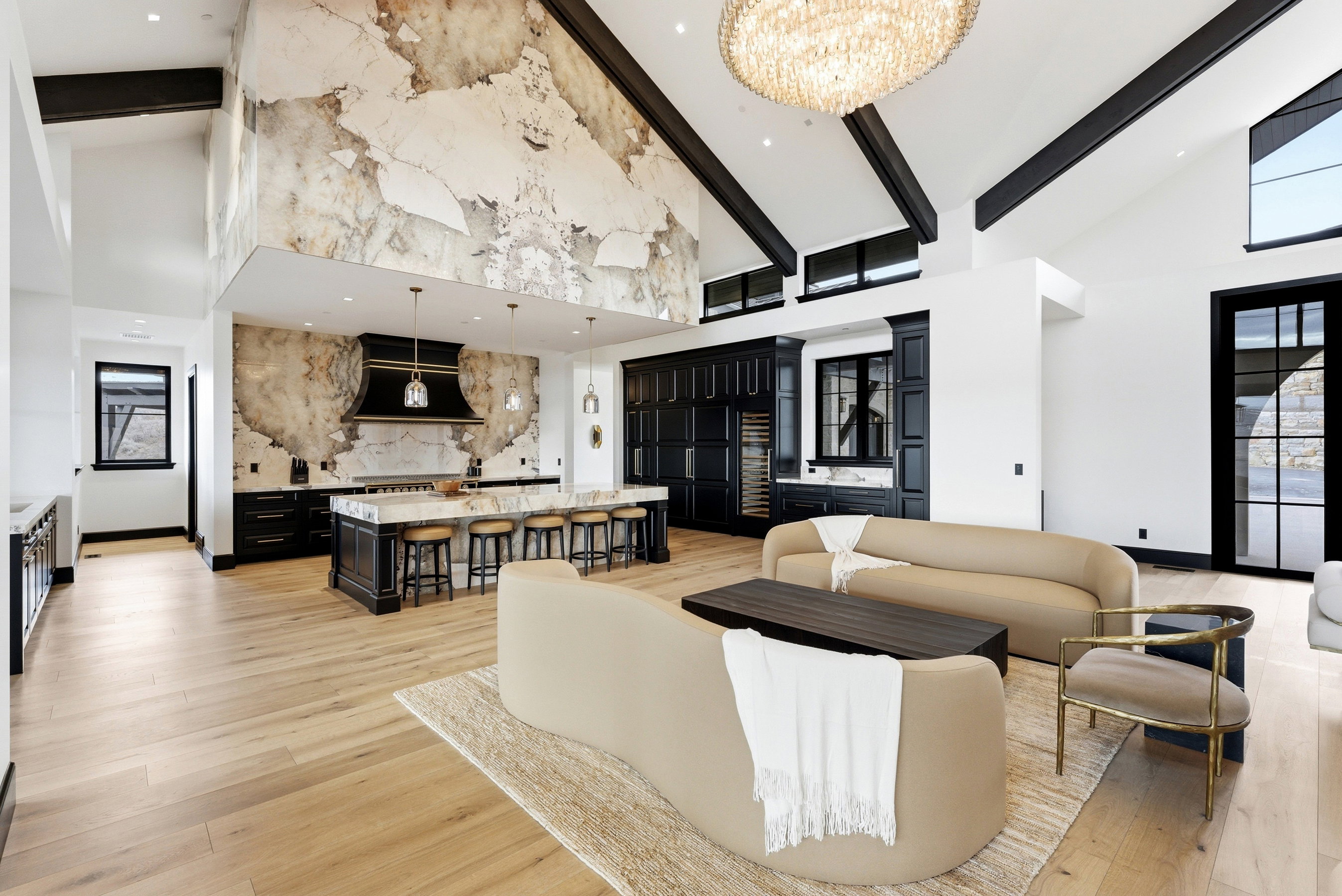 Open-concept living space featuring vaulted ceilings with exposed black beams, light wood-finish flooring, and a large crystal chandelier