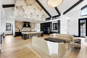 Open-concept living space featuring vaulted ceilings with exposed black beams, light wood-finish flooring, and a large crystal chandelier