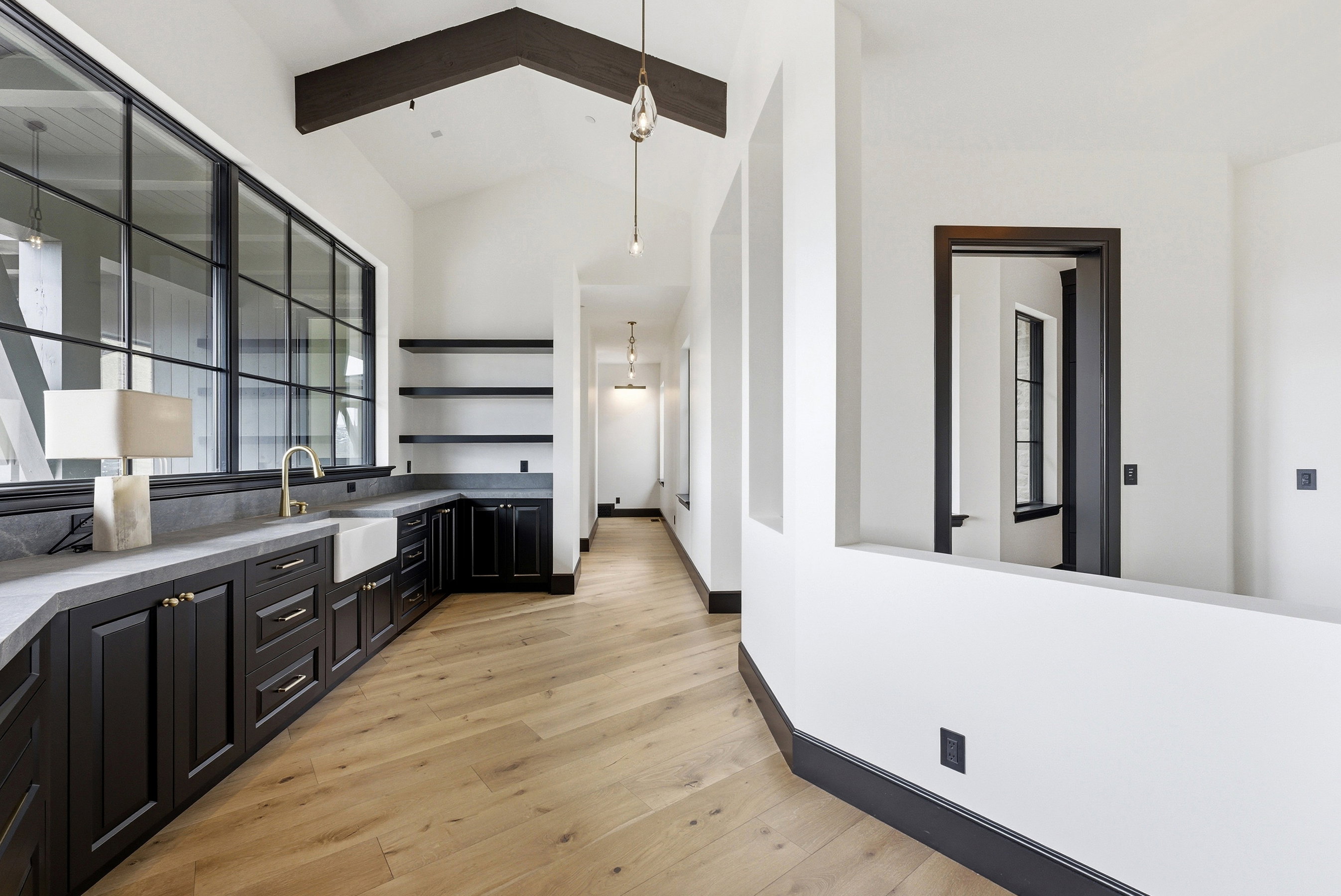 Expansive multi-paned windows illuminate a modern wet bar featuring a farmhouse sink, brass faucet, and dark custom cabinetry with stone countertops